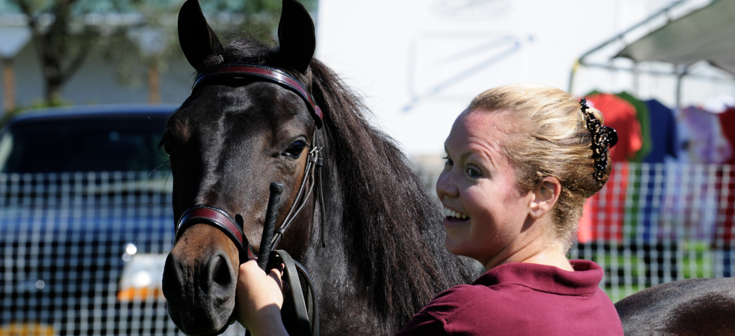 A woman with blonde hair in a bun smiling and holding the bridle of a dark brown horse with a black mane, at an outdoor event with colorful tents and a fence in the background.