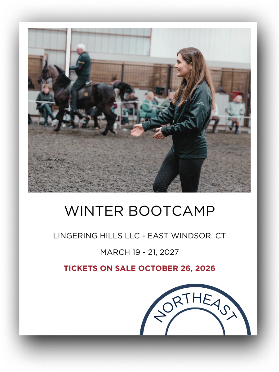 A woman at an indoor riding arena, clapping and smiling while horses and riders appear in the background during a winter bootcamp event.