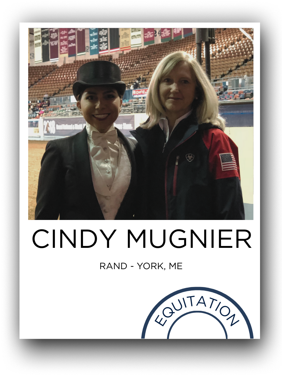 Two women standing together at a horse arena, one dressed in a black equestrian jacket with an American flag patch, and the other in a white show outfit with a black riding helmet, with empty stadium seats and banners in the background.