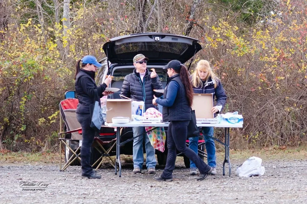 Group of five women gathered around a table outdoors during fall, with a black SUV and trees with colorful leaves behind them, having a picnic or outdoor meal.