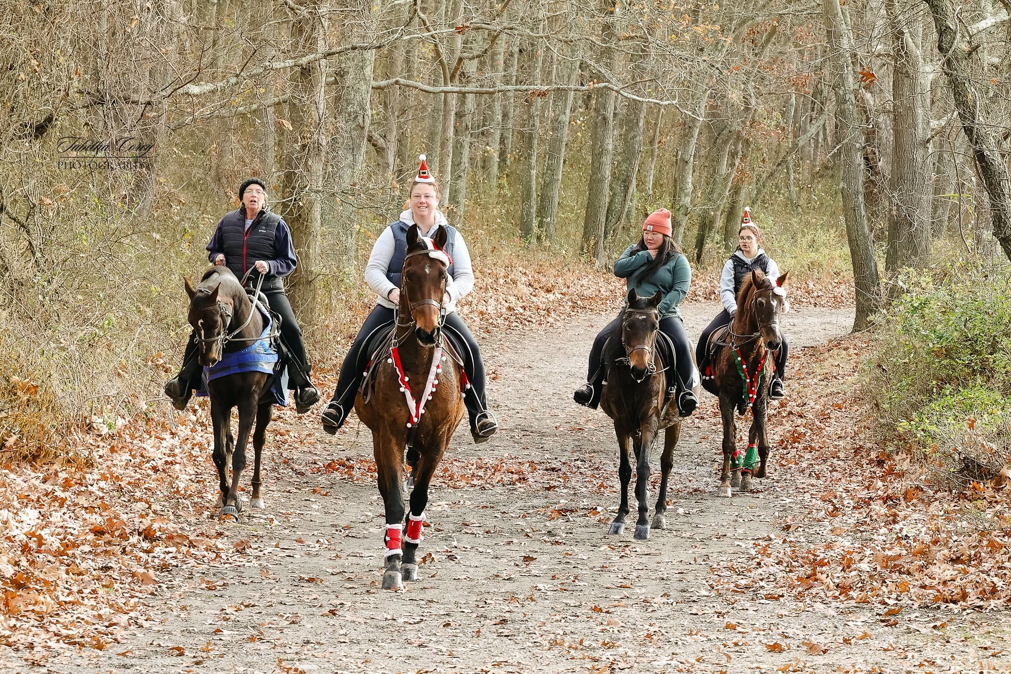 Four women riding horses on a wooded trail decorated for Christmas, with some wearing festive accessories.