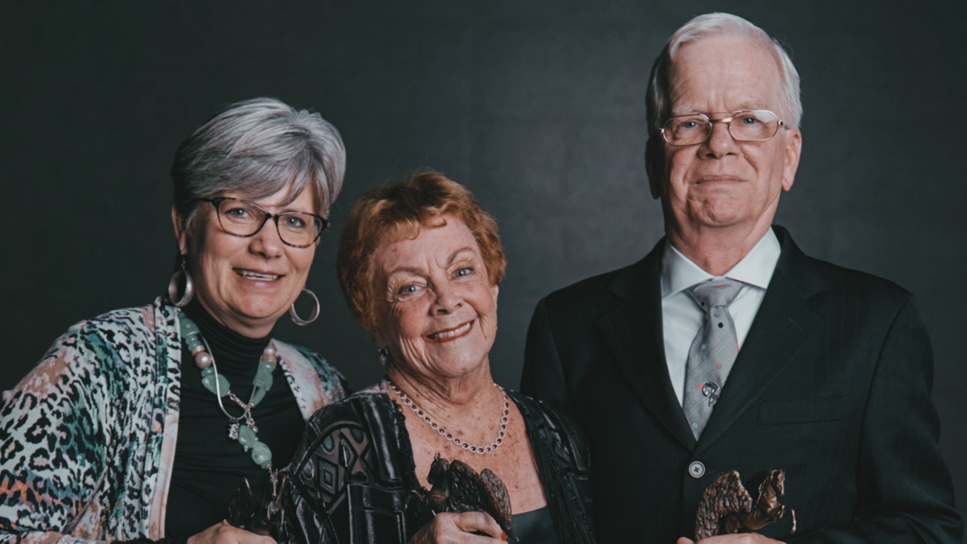 Portrait of three elderly adults, two women and one man, standing together and smiling, dressed in formal attire against a dark background.