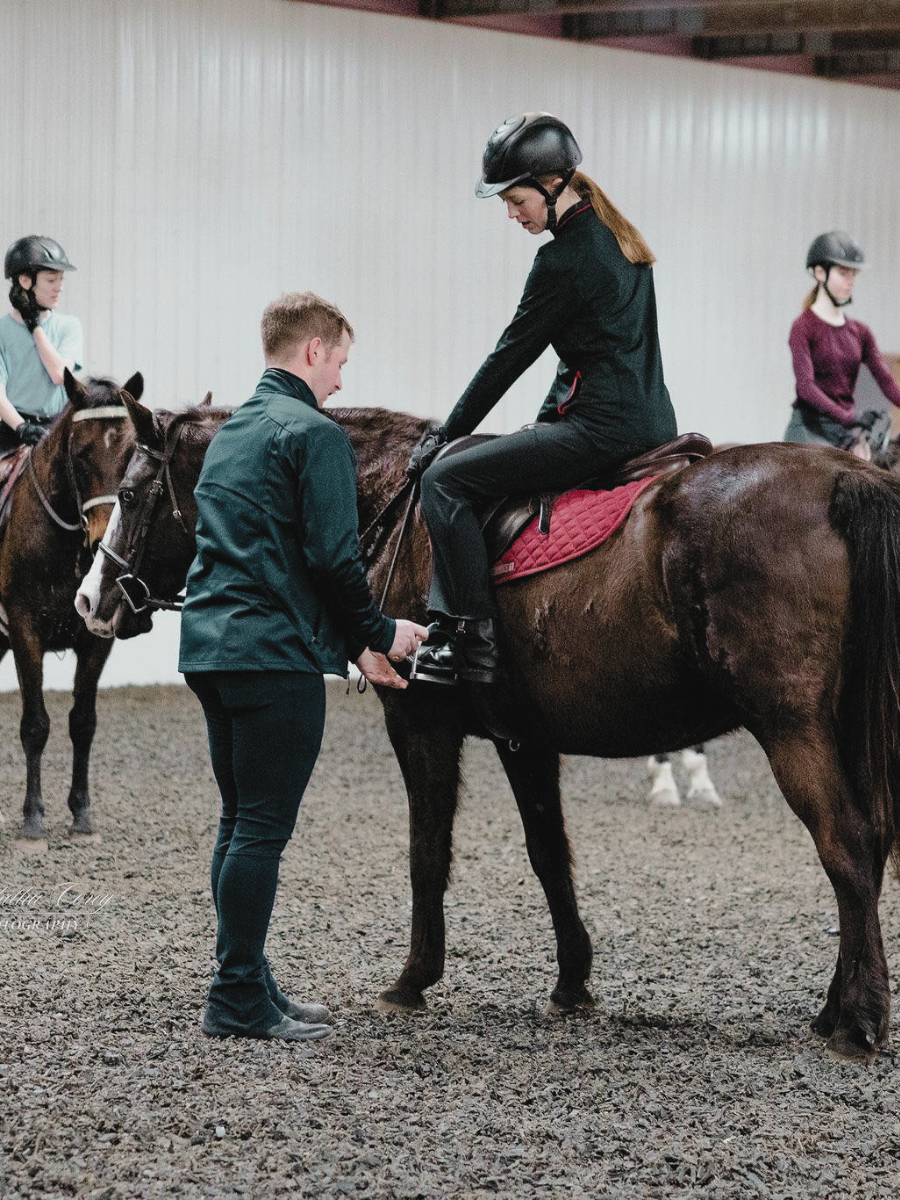 A woman in riding gear sitting on a brown horse while a man in black assist her in tying her riding boots inside an indoor riding arena. Other riders on horses are visible in the background.