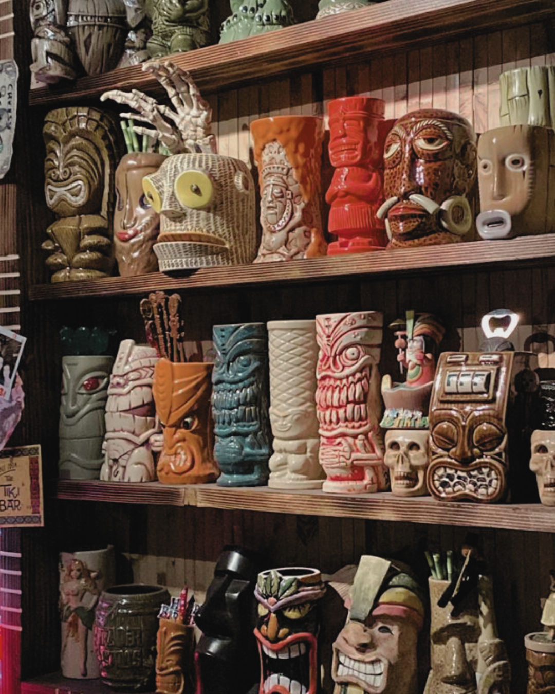 A shelf display of various colorful Tiki mugs and masks, featuring Polynesian and tribal-inspired designs.