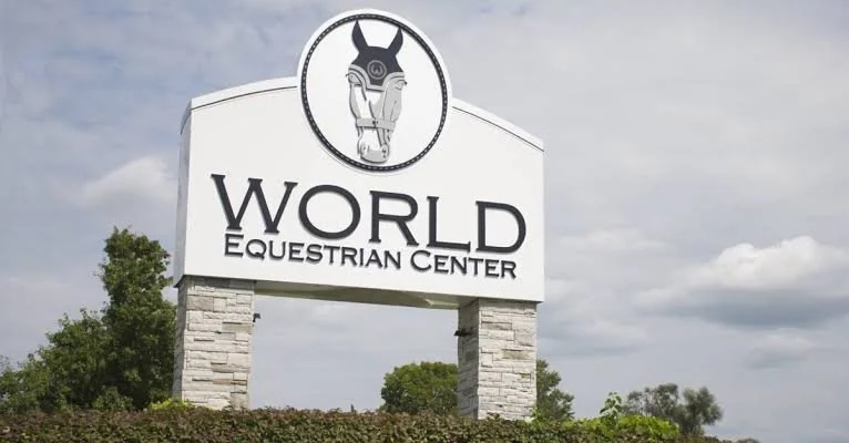 Sign for the World Equestrian Center with a horse's head logo at the top, set against a cloudy sky and surrounded by greenery.