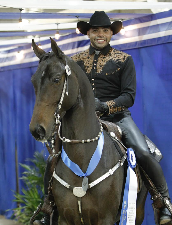 Smiling man wearing a cowboy hat and western shirt riding a horse with medals and ribbons, blue curtains in the background.