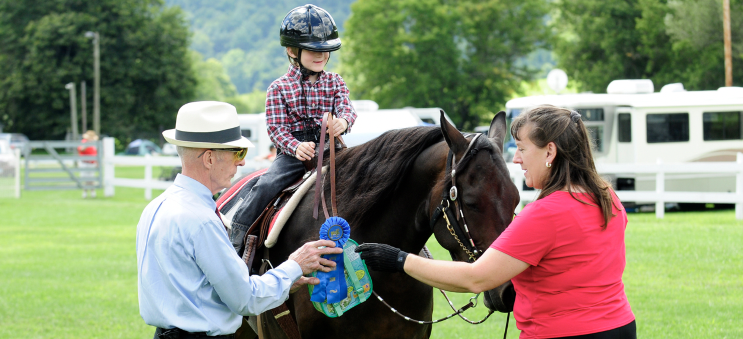A young girl wearing a helmet sits on a dark brown horse during an outdoor daytime event. An older man in a light blue shirt and white hat, holding a ribbon, and a woman in a pink shirt, stand beside the horse. The background features green trees, tr