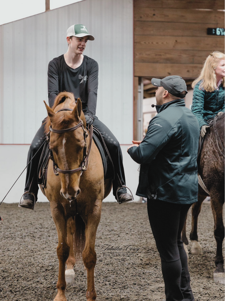 A young person wearing a gray and green baseball cap and a black long-sleeve shirt is sitting on a brown horse and talking to a man in black jacket and cap, inside an indoor riding arena with wood and metal walls.