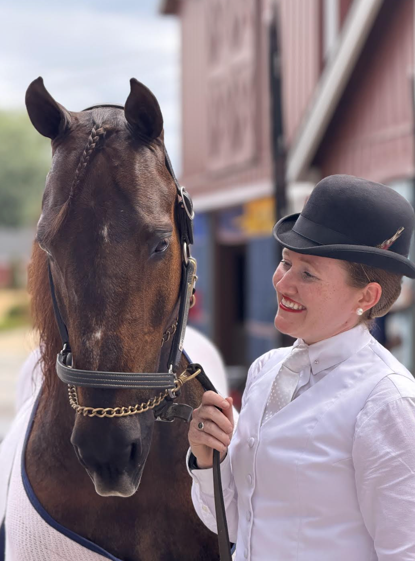 A woman in a white shirt and black hat smiling while holding a horse's bridle outside a barn.
