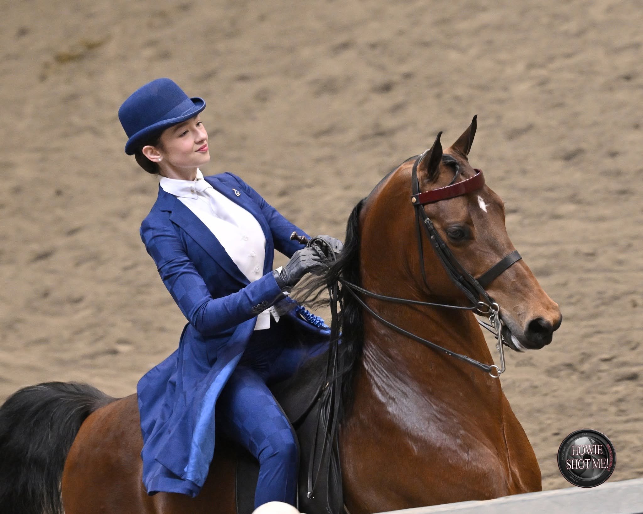 A woman dressed in a blue blazer, white shirt, and a blue hat riding a brown horse on an indoor riding arena.