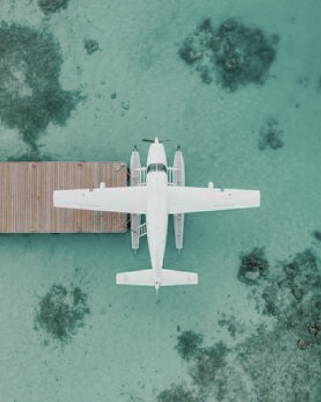 Top-down view of a white seaplane docked at a wooden pier on clear, shallow water with visible rocks.