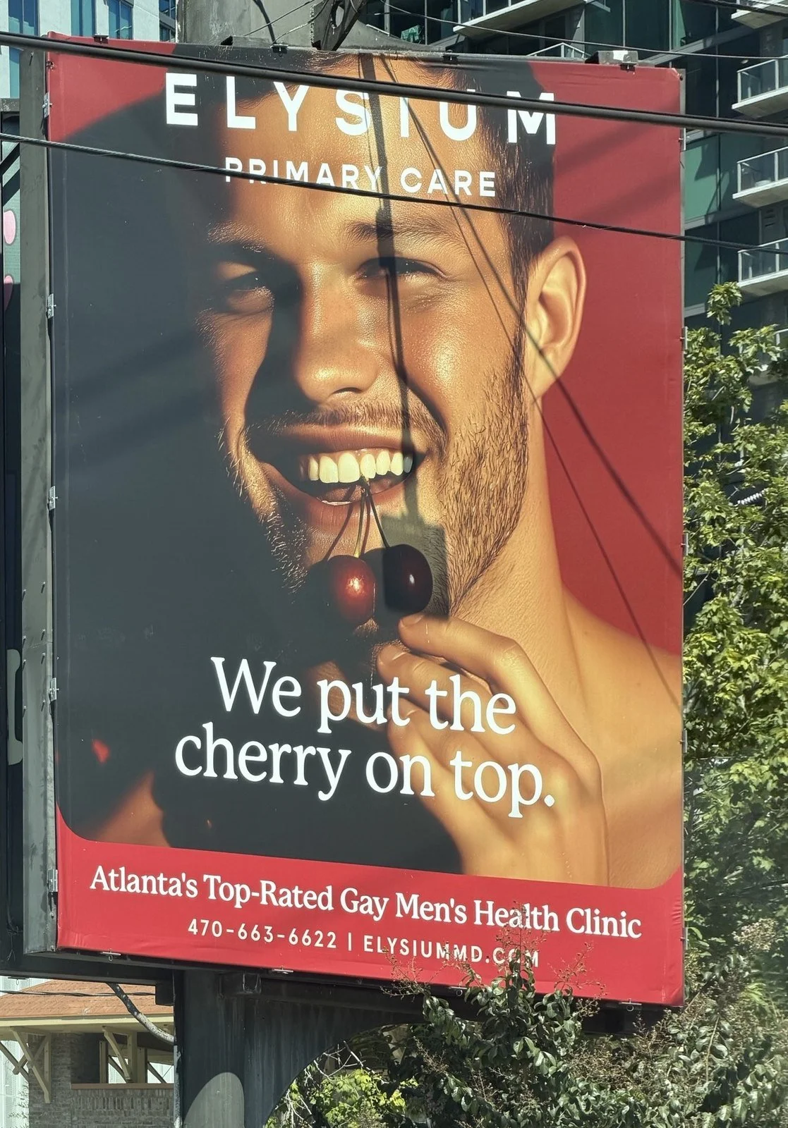 Elysium Primary Care billboard in Atlanta: a smiling man holding two cherries near his mouth, headline "We put the cherry on top." and the line "Atlanta's Top-Rated Gay Men's Health Clinic."