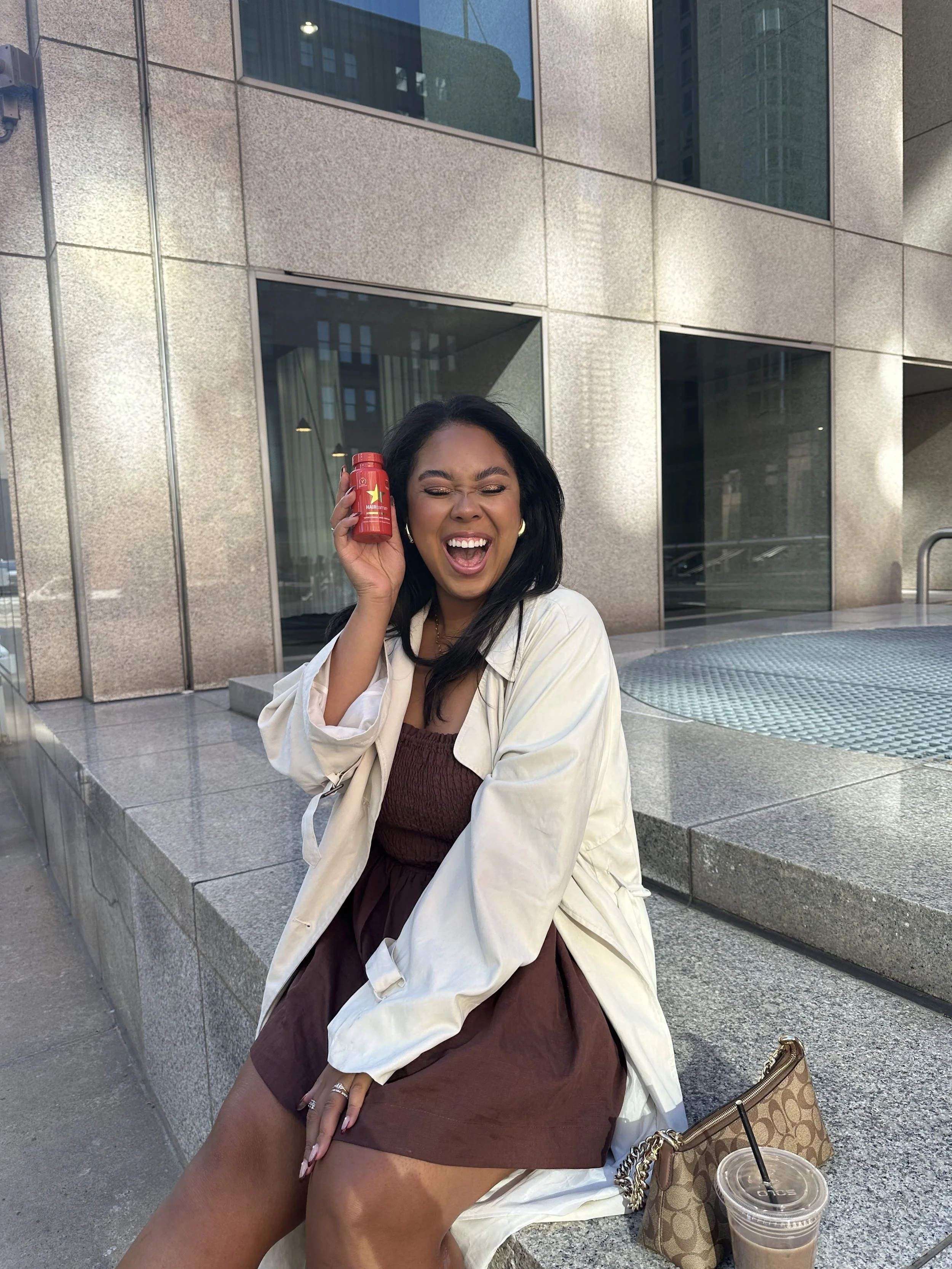 A woman sitting on a concrete ledge outside a building, holding a red bottle near her ear, smiling with her eyes closed. She is wearing a brown dress and a white jacket, with a purse and iced coffee placed beside her.