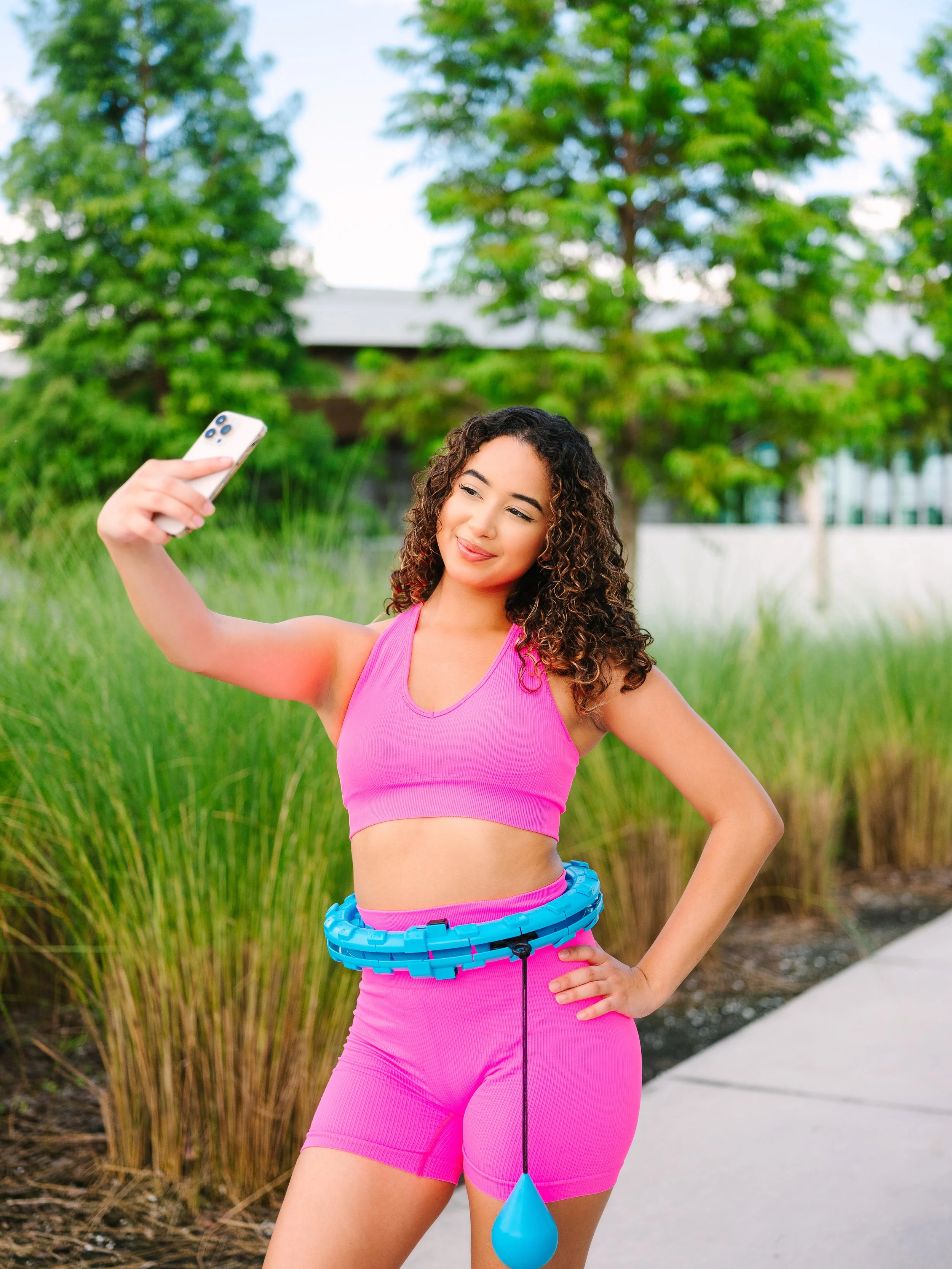 Young woman in pink workout clothes taking a selfie outdoors with greenery and trees in the background.