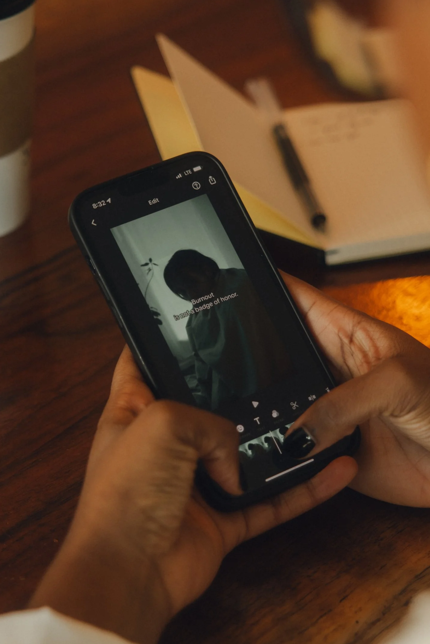 A person holding a smartphone displaying an image of a child with the caption 'Burnout is not a badge of honor.' The phone is on a wooden surface with an open notebook and a pen in the background.