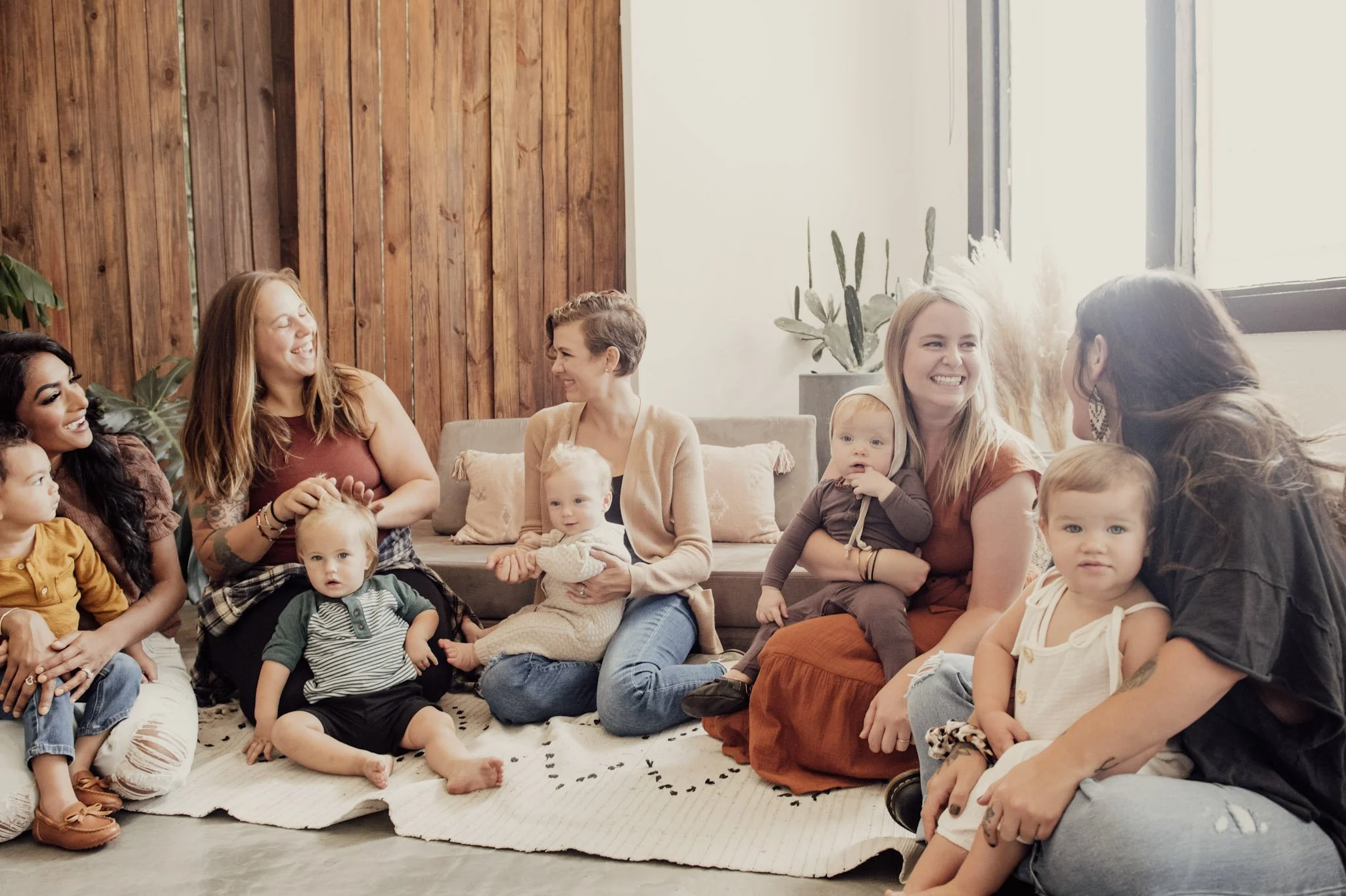 A group of women and children sitting on a rug in a room with wooden paneling and large windows, smiling and engaging with each other.