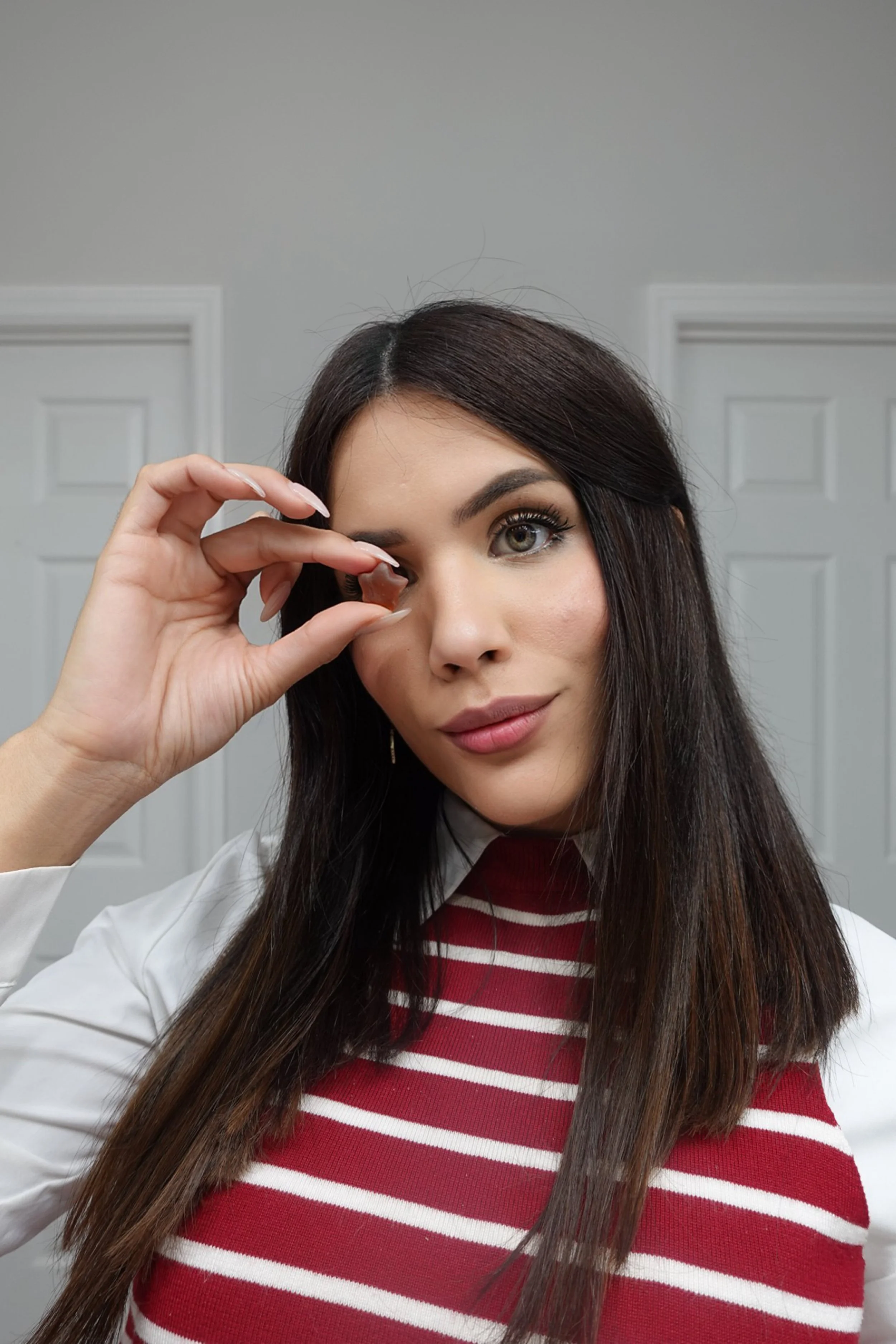 A woman with long dark hair holding a small object near her eye, inside a room with white paneled walls and doors.