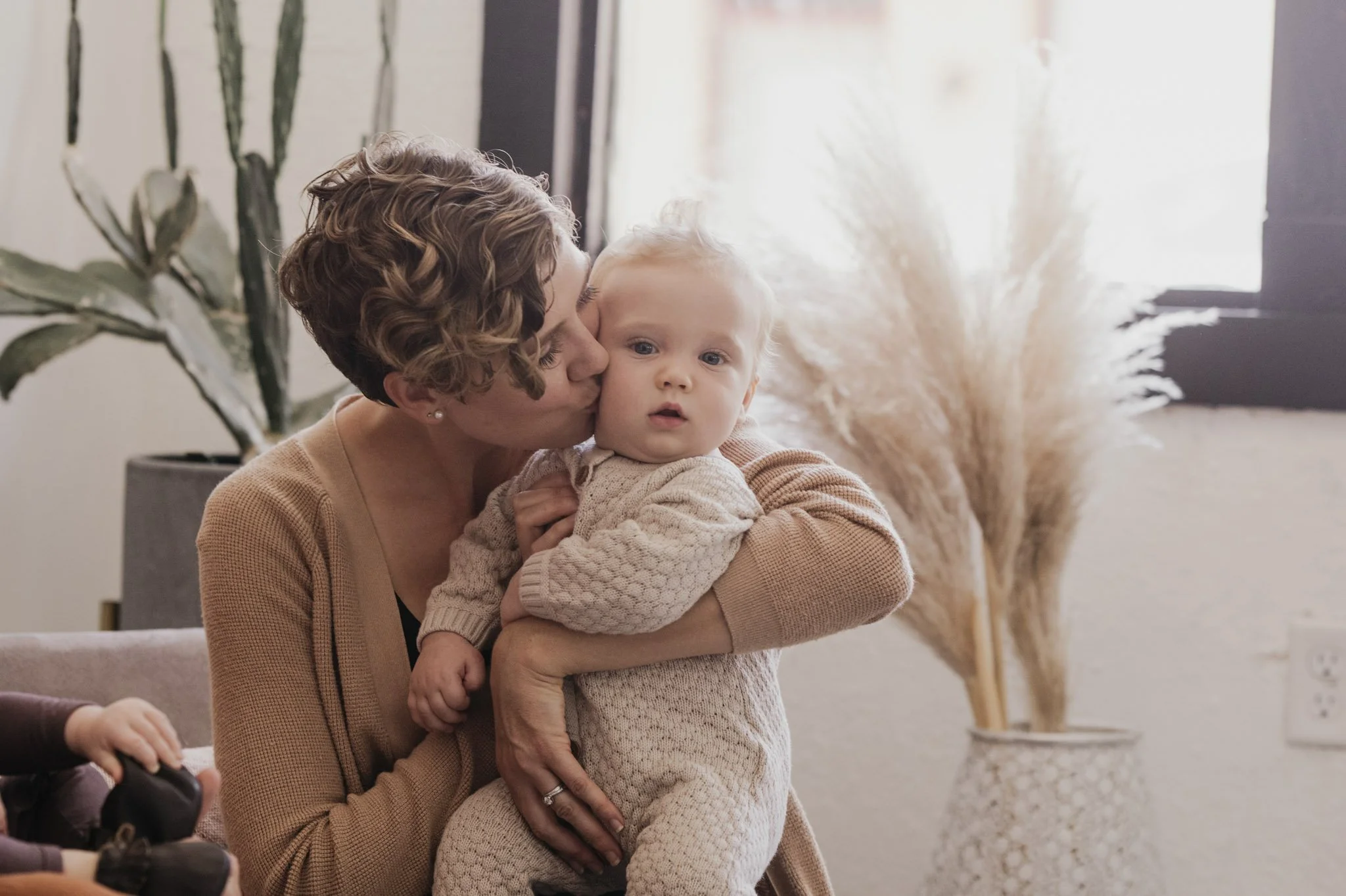 A woman with short curly hair is kissing a young child on the cheek. The child has blonde hair and wide eyes, wearing a cozy sweater. They are inside a room with a large plant and pampas grass in the background.