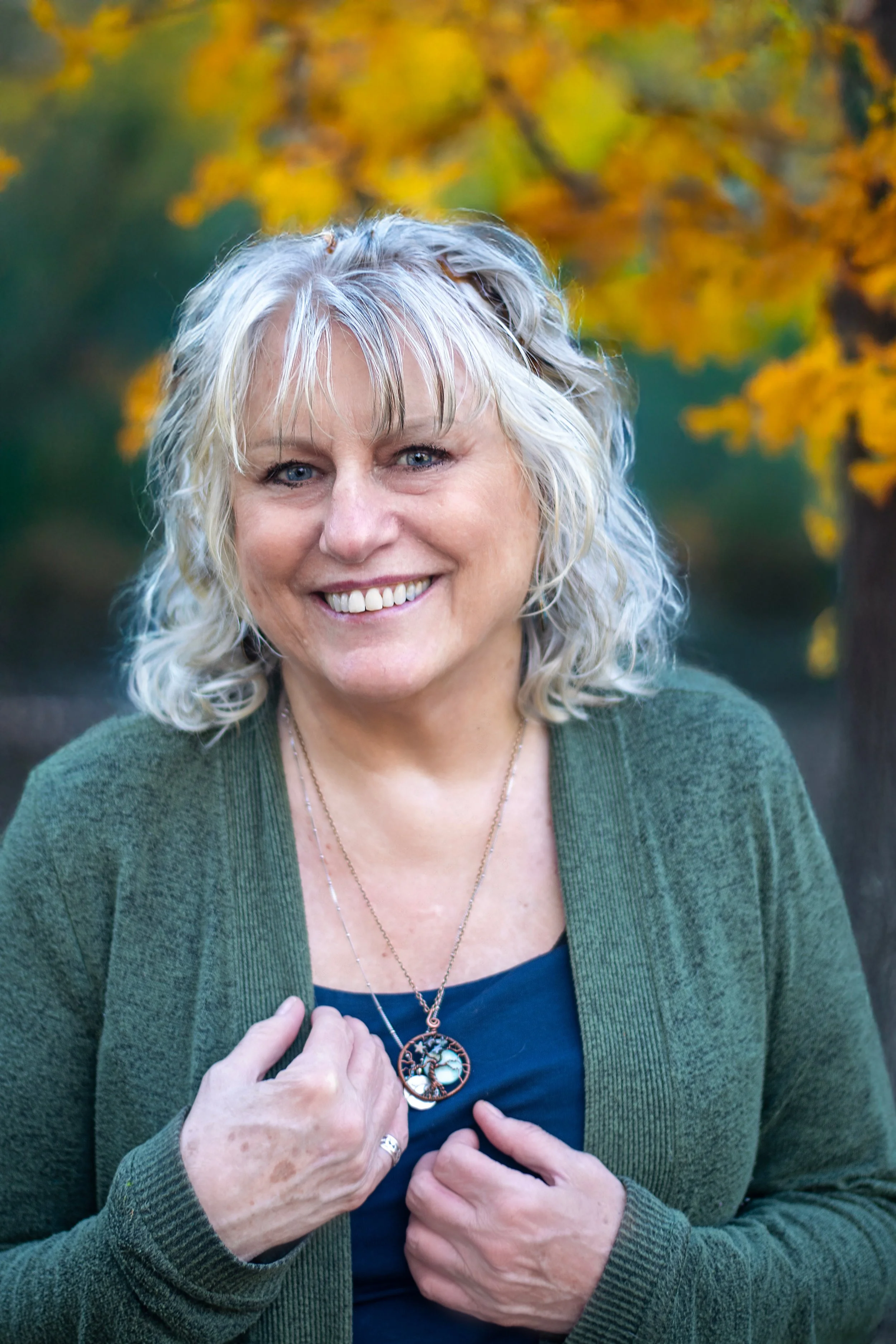 A smiling woman with curly gray hair, wearing a green cardigan over a black top, standing outside with yellow autumn leaves in the background.