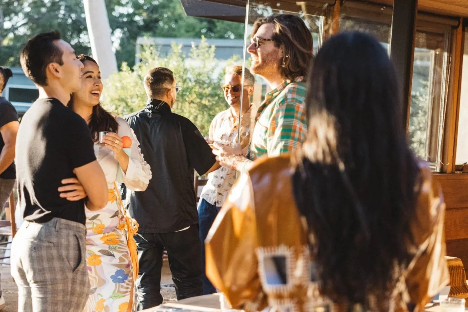 People socializing outdoors at a casual gathering, some standing and chatting, others smiling and laughing, with trees and a building in the background and a table with dishes in the foreground.