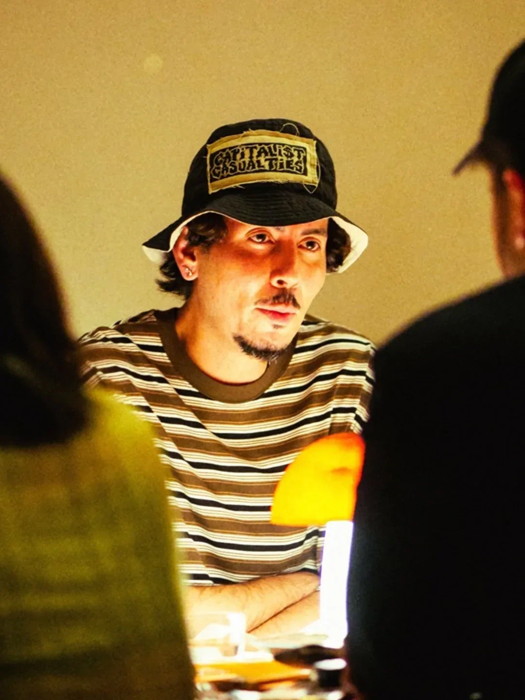 A man with a goatee wearing a black bucket hat with a patch that says 'Capitalist Casualties' and a striped shirt in a dimly lit room, seen from the front with others partially visible in the foreground.