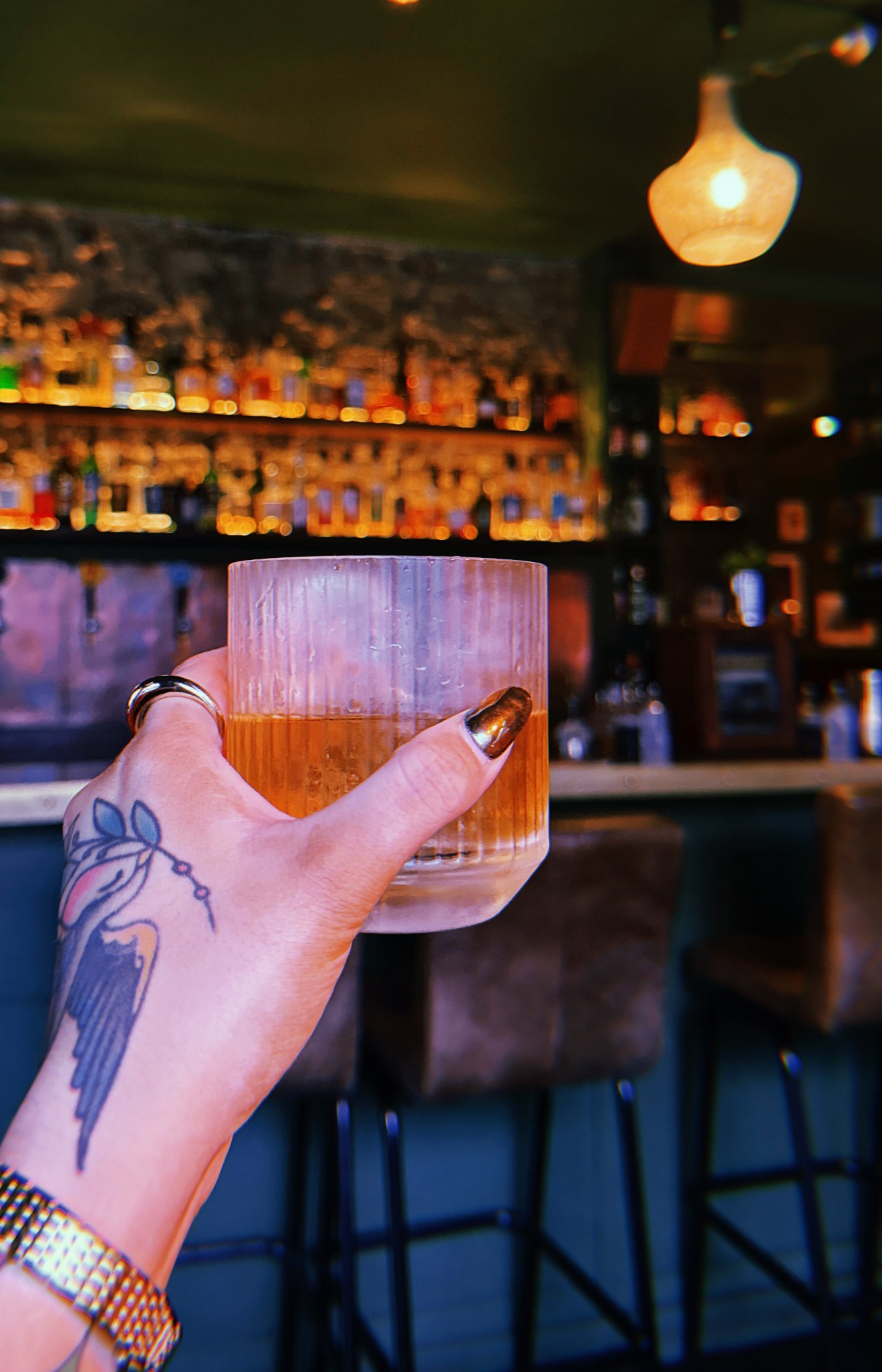 A hand with a tattoo of a bird and flowers holding a glass of amber-colored drink inside a bar, with shelves of bottles and a warm hanging light in the background.