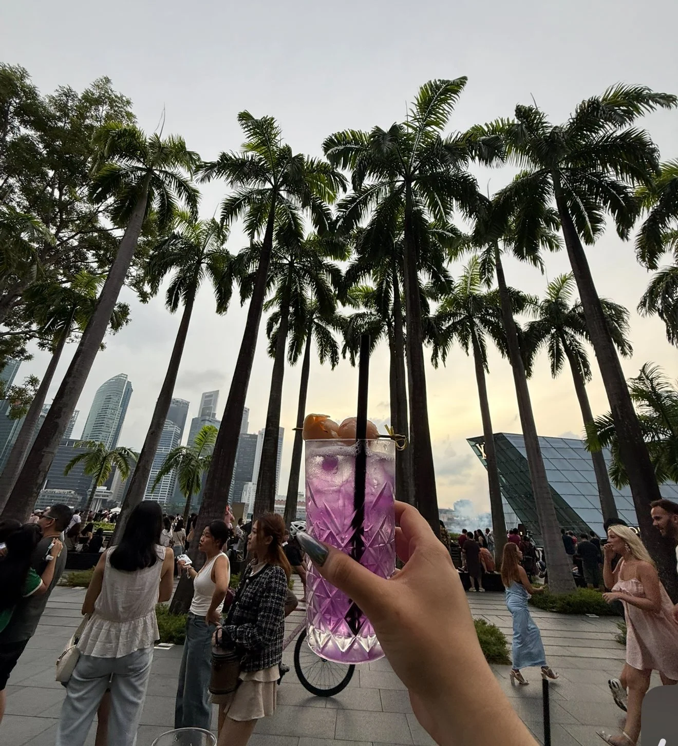 A hand holding a tall glass of purple cocktail with ice and garnish in a Singapore park with palm trees, skyscrapers, and a crowd of people in the background.