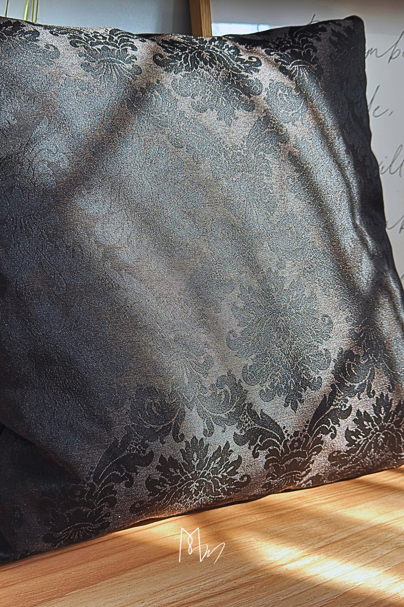 Un coussin noir avec un motif damassé argenté, placé sur une surface en bois