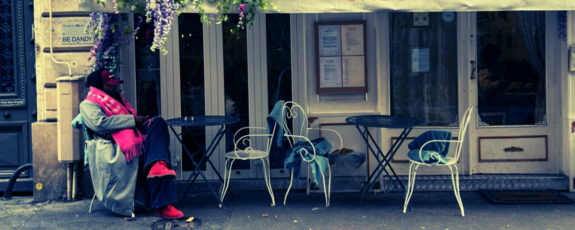 Une personne assise à une table d'extérieur devant un restaurant ou café, portant des vêtements gris et un foulard rose, avec des chapeaux et vêtements rouges, sur un trottoir.