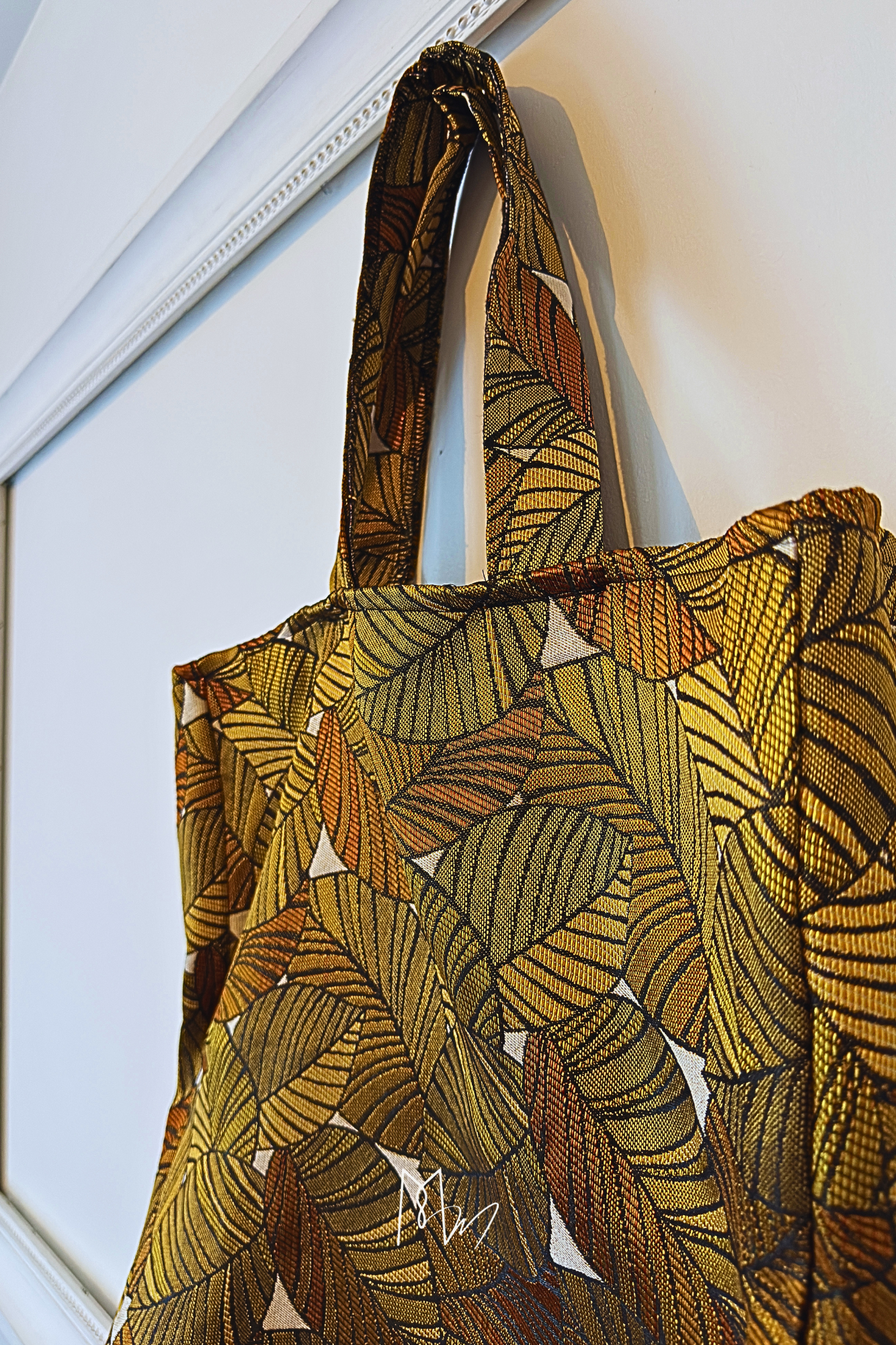 Sac en tissu avec motif de feuilles colorées en tons de jaune, orange et vert, suspendu à une poignée de porte.