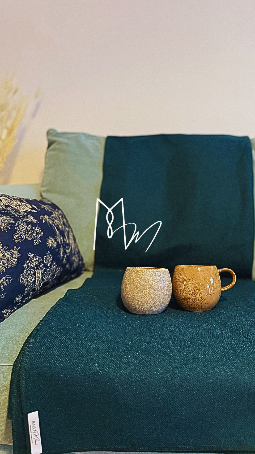 Deux tasses en céramique beige posées sur une banquette en tissu vert foncé, avec des coussins gris clair, vert clair et un coussin avec un motif floral blanc et bleu foncé en arrière-plan.