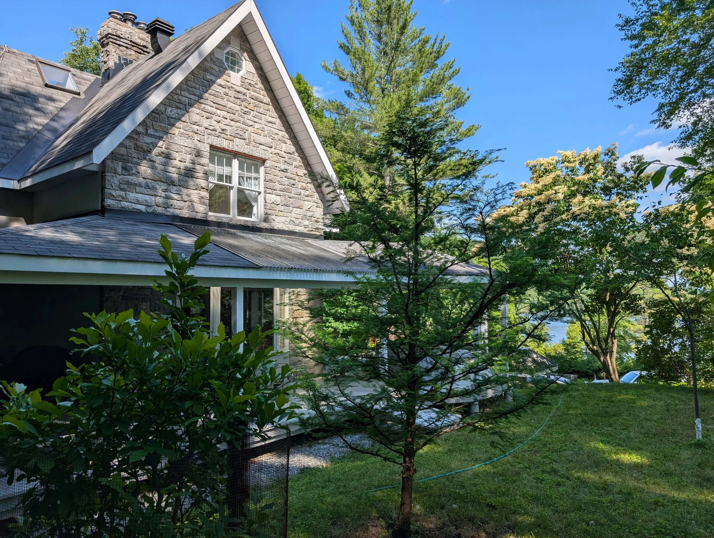 A stone house with a gabled roof, surrounded by green trees and a grassy yard under a clear blue sky.