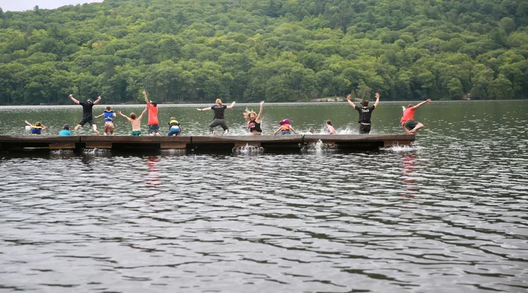 People jumping off a wooden dock into a lake with a forested hill in the background.