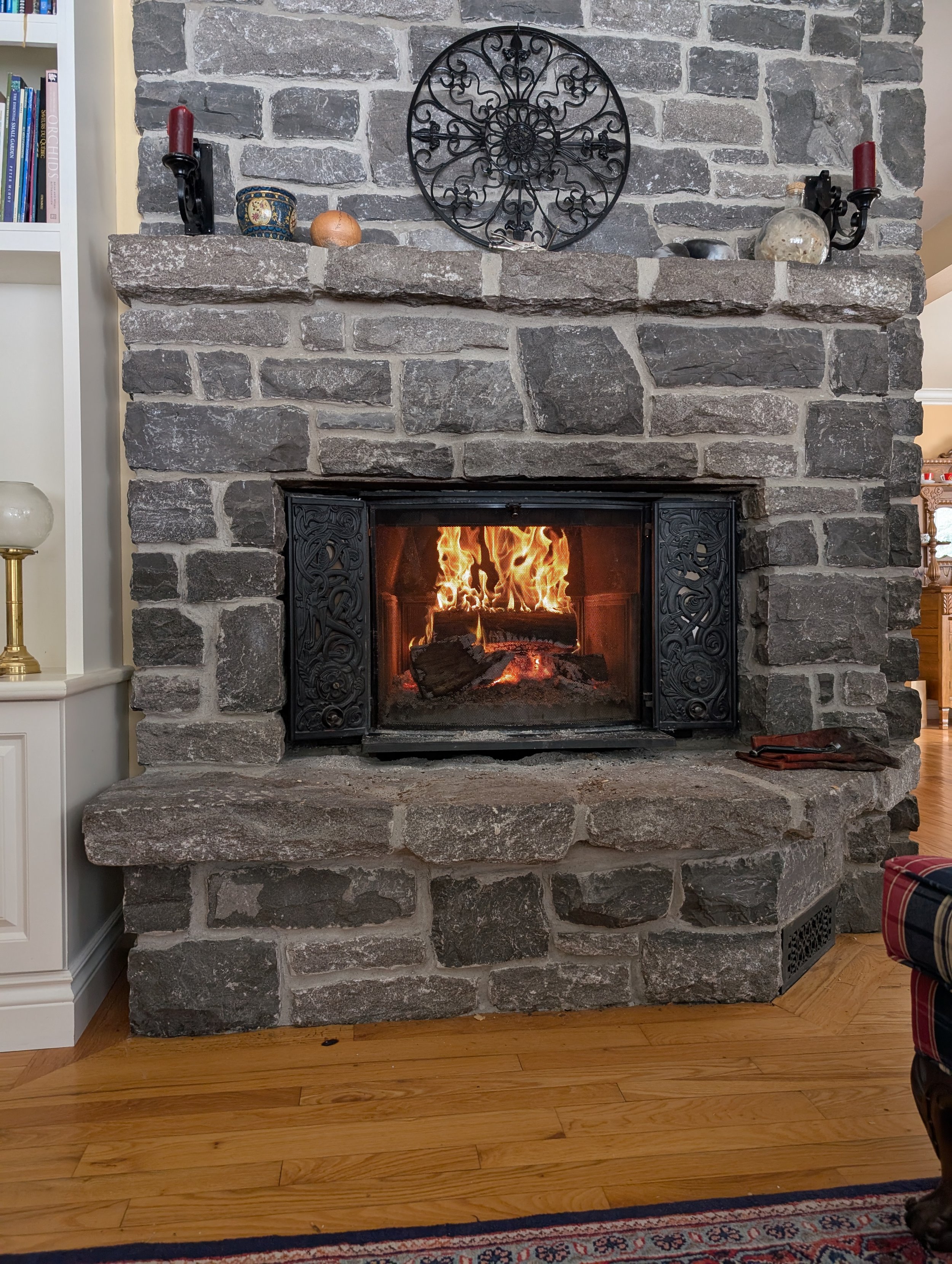 A stone fireplace with a fire burning inside, decorated with black wrought iron embellishments on the frame and a decorative metal wall piece above. The mantle holds candles and small decorative objects. To the left, part of a white bookshelf is visi