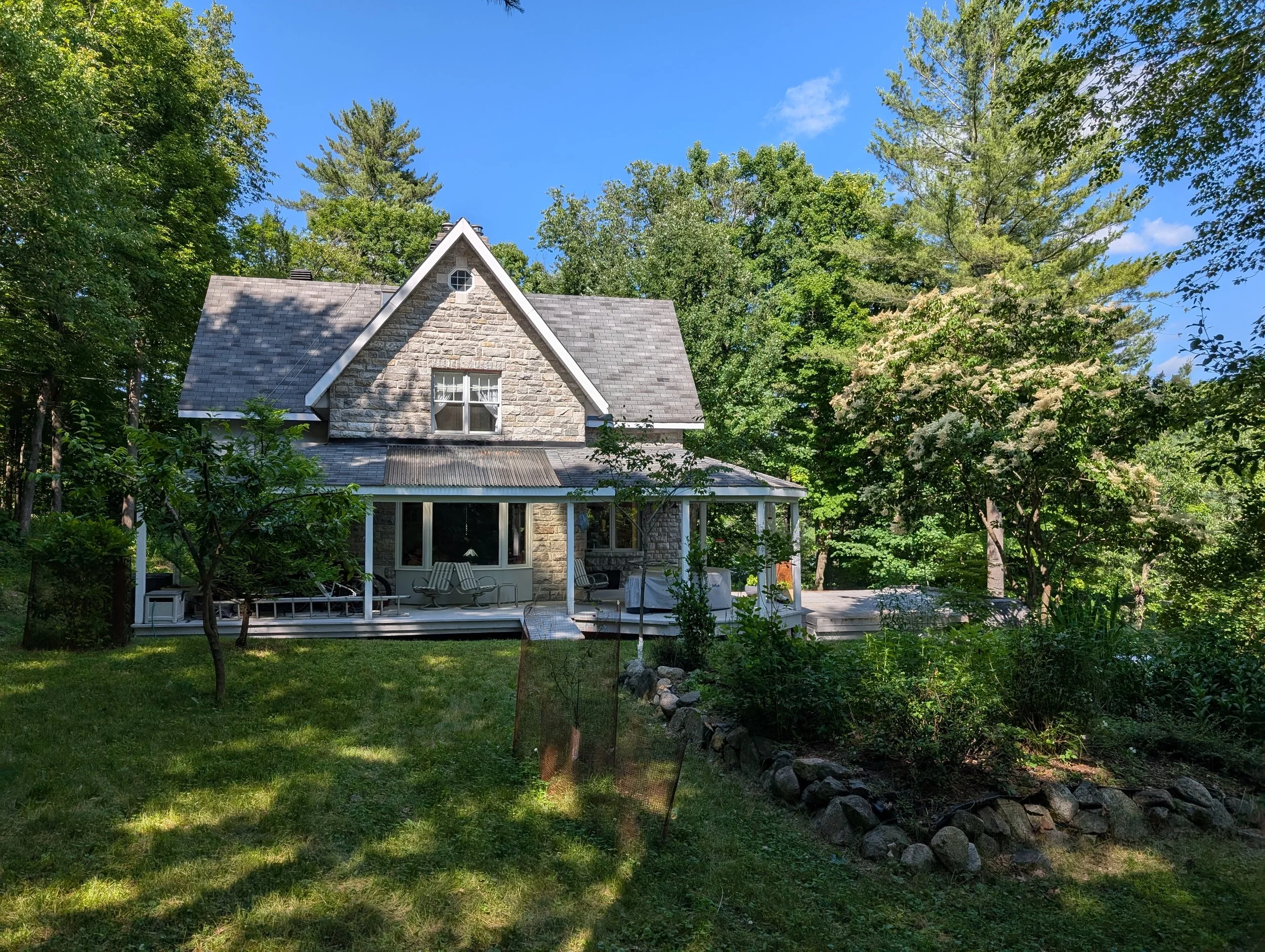 A stone house with a large front porch surrounded by trees and greenery under a clear blue sky.