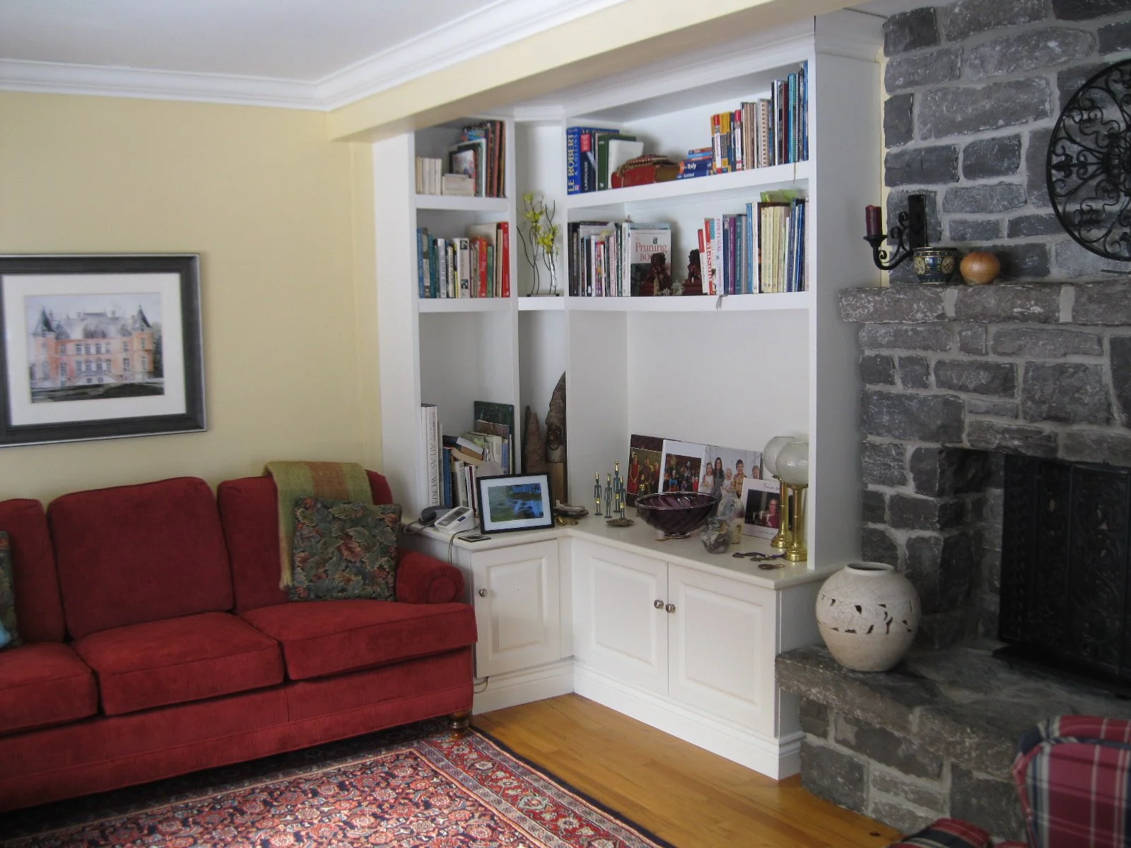 Living room with a red sofa, a built-in white bookshelf filled with books, a framed watercolor painting on a yellow wall, a stone fireplace with decorative items, and a wooden floor with an ornate rug.
