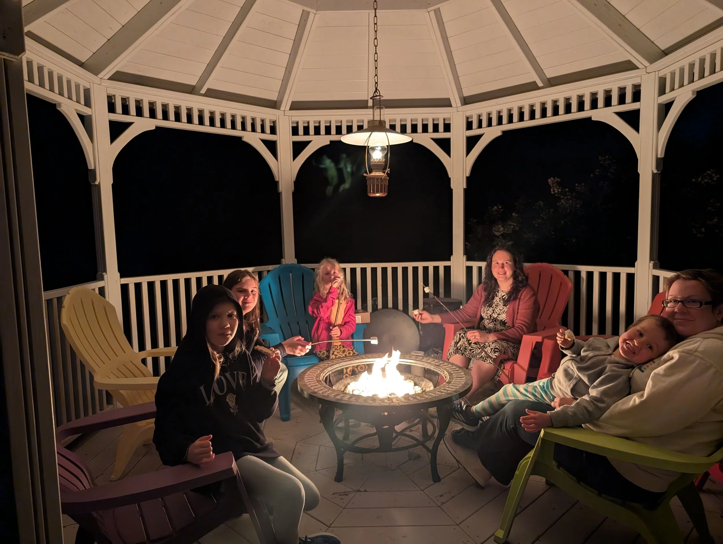Group of six people sitting around a lit outdoor fire pit in a gazebo at night, with chairs and a hanging lantern overhead.