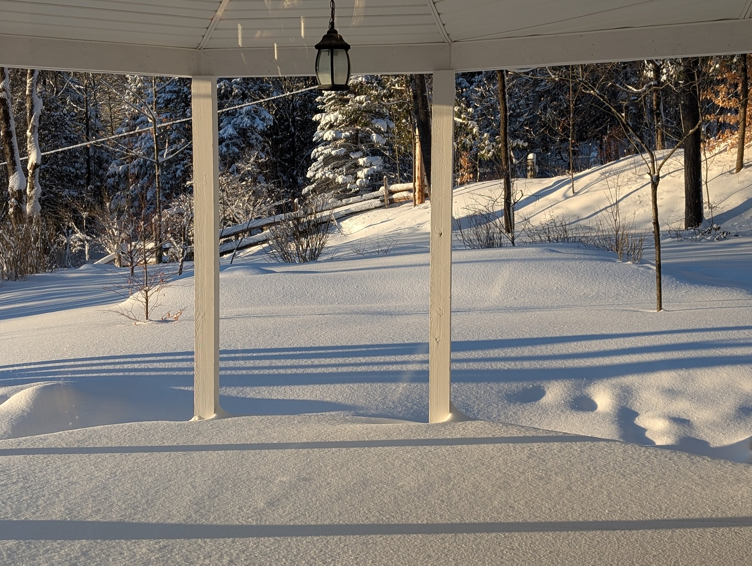 Snow-covered backyard seen from porch with vertical support posts and a hanging lantern, trees and bushes in the background under a clear sky.