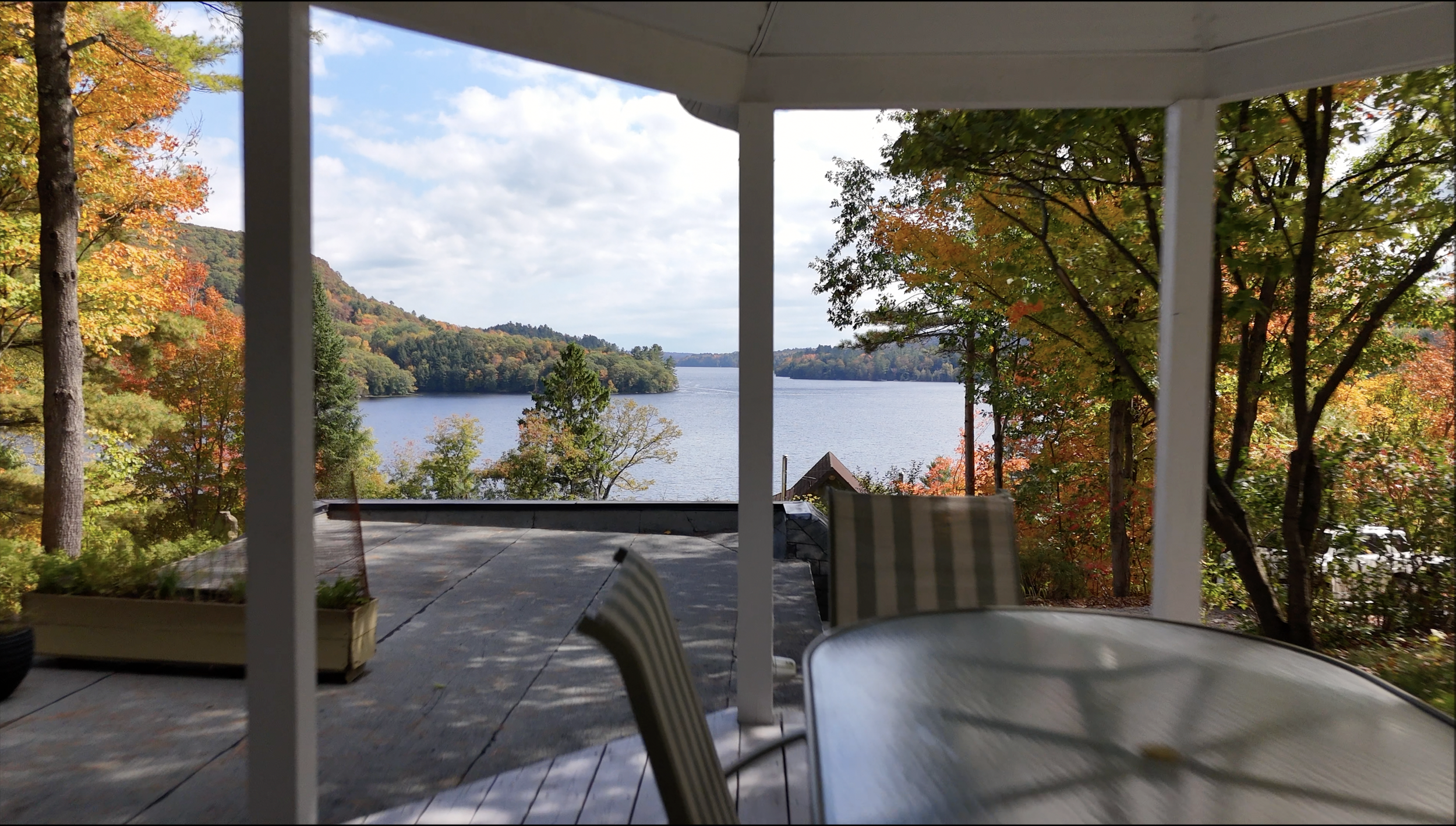 View of a lake surrounded by autumn-colored trees, seen from a covered porch with a patio table and chairs.
