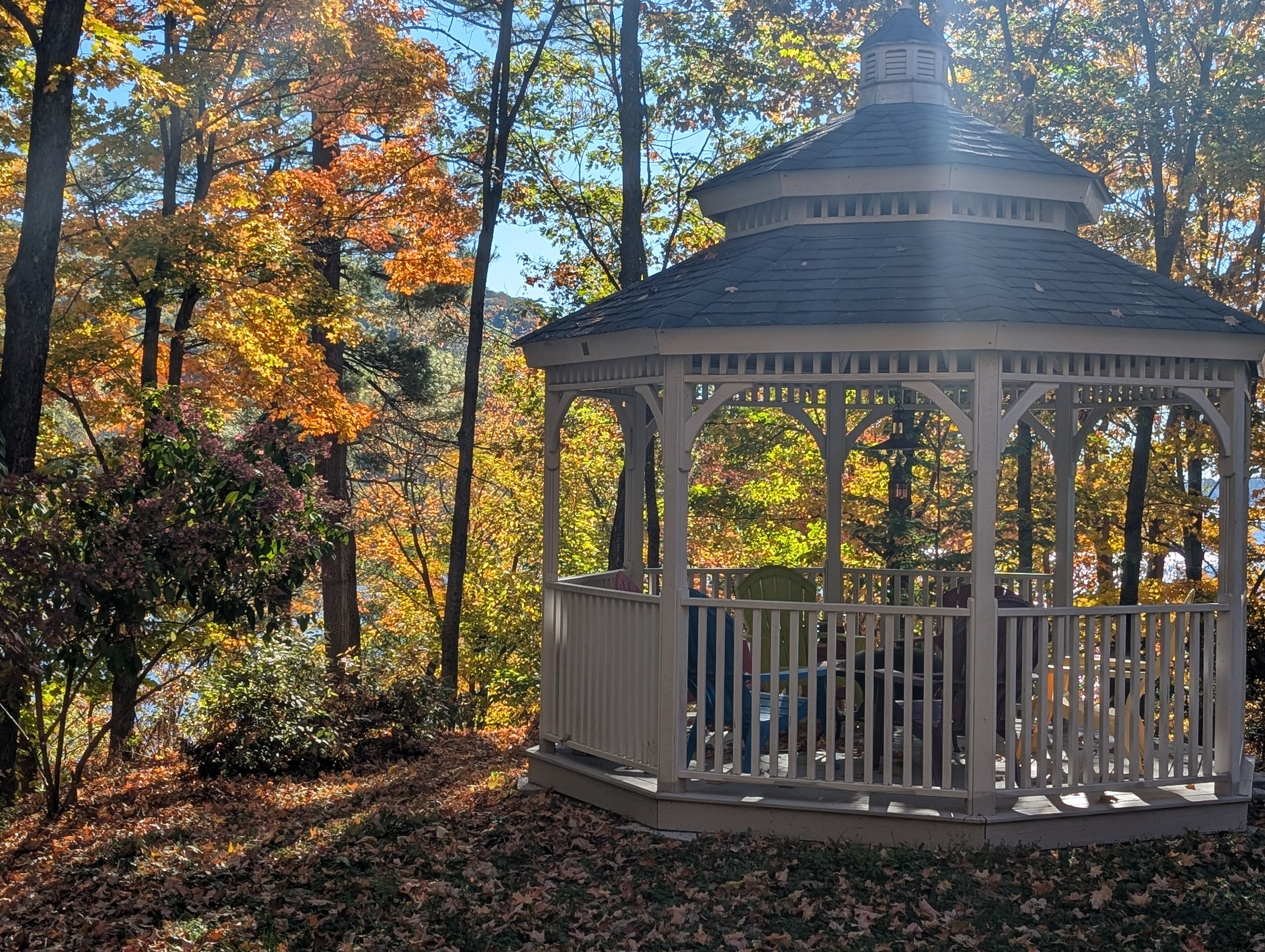 A white wooden gazebo in a wooded area during fall, with colorful autumn leaves on trees and on the ground.