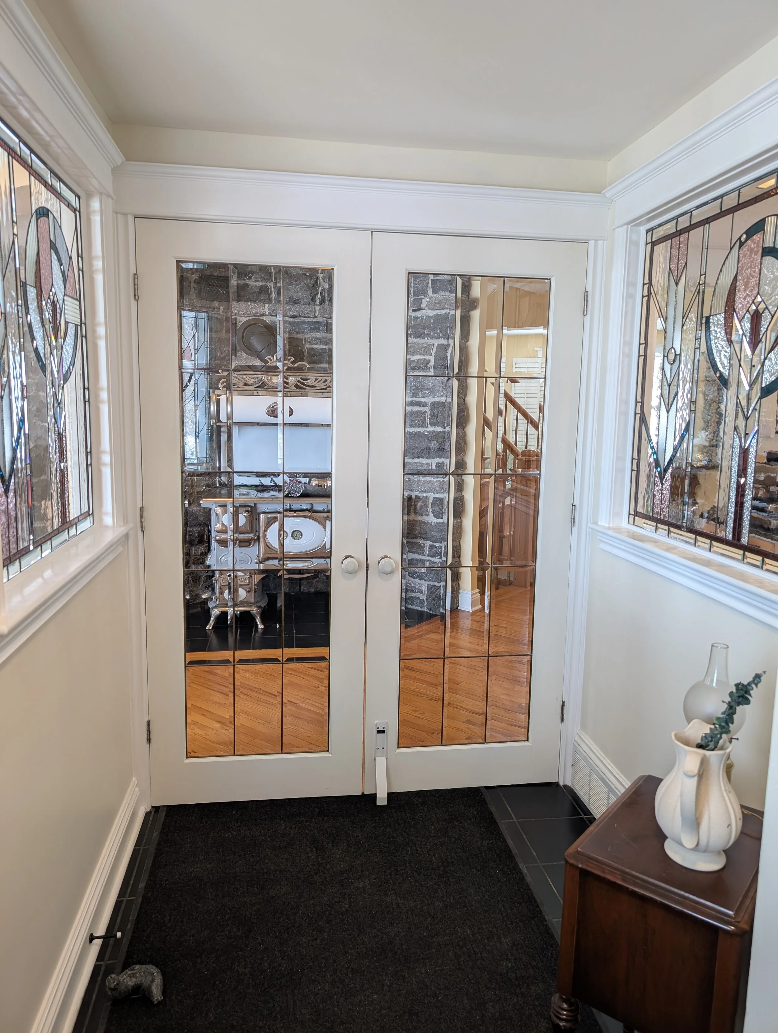 Interior view of a foyer with glass-paned double doors leading to a kitchen, dark floor mat, decorative stained glass windows, and a small wooden table with vases and greenery.