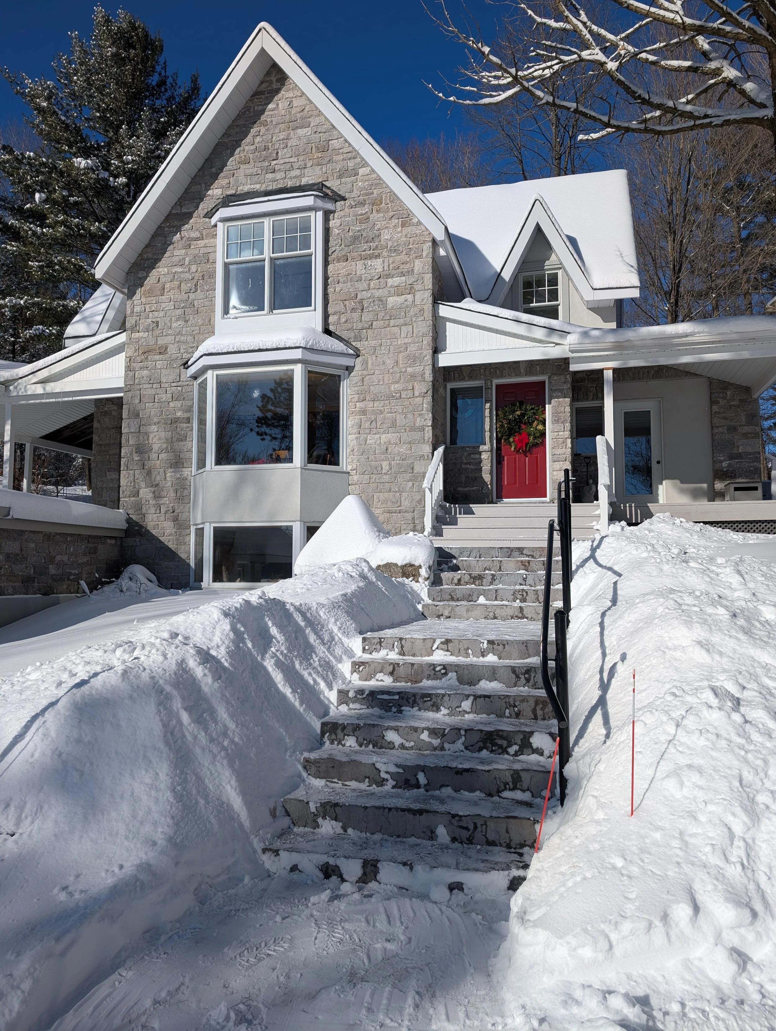 Snow-covered front steps leading to a house with a red door, stone and white siding exterior, large bay windows, and surrounded by snow and trees.