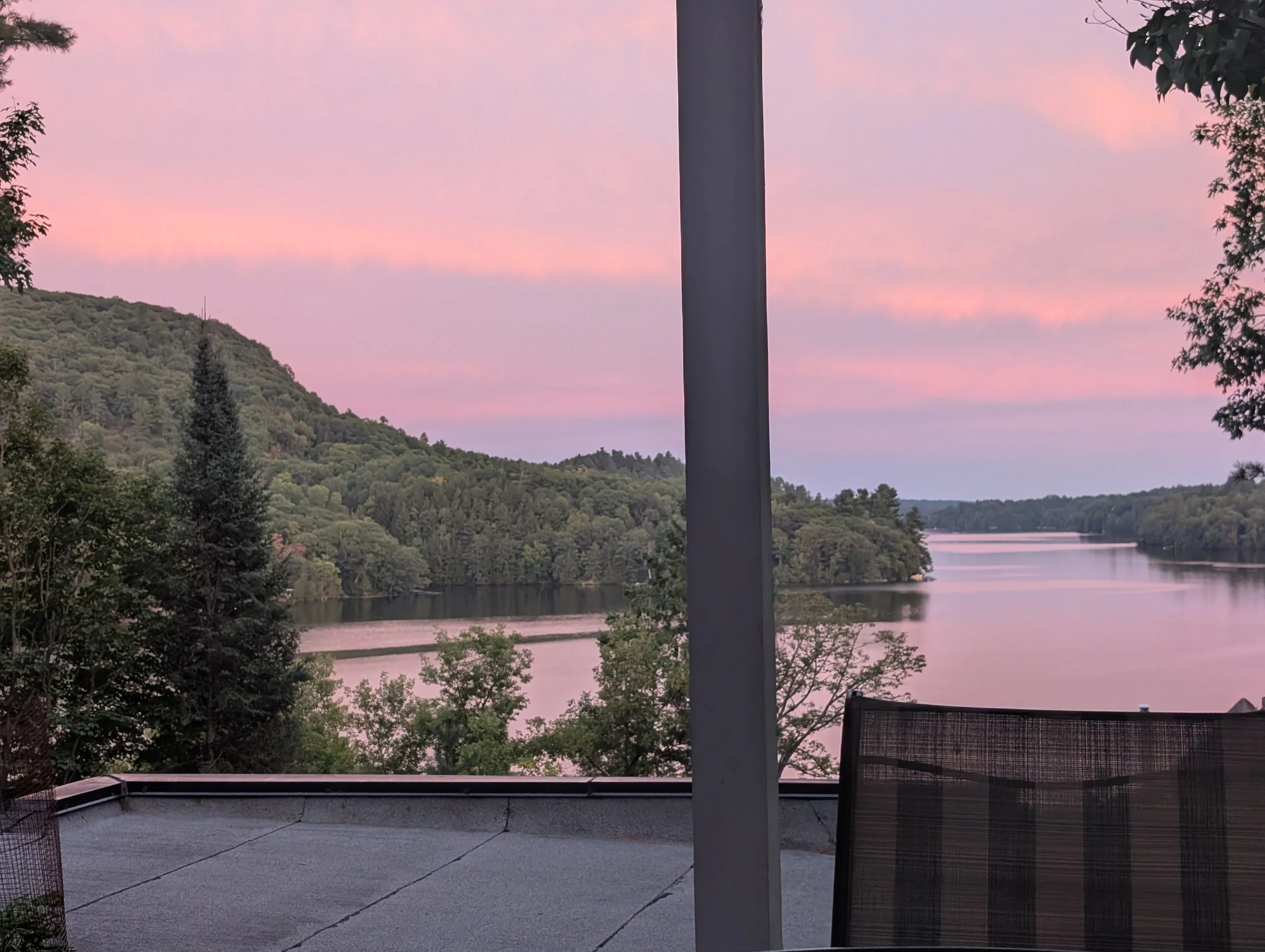 View of a river surrounded by green trees and hills during sunset, seen from a rooftop with patio furniture in the foreground.