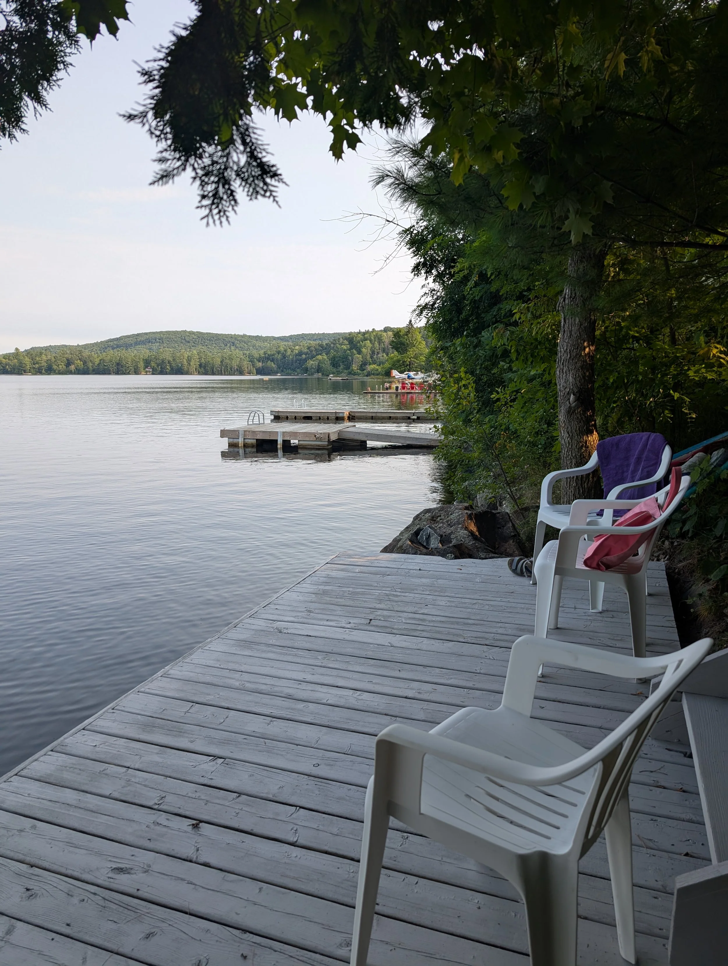 A lakeside wooden dock with white plastic chairs, some with towels draped over them, overlooking a calm lake with a small floating dock and forested hills in the background, shaded by trees.