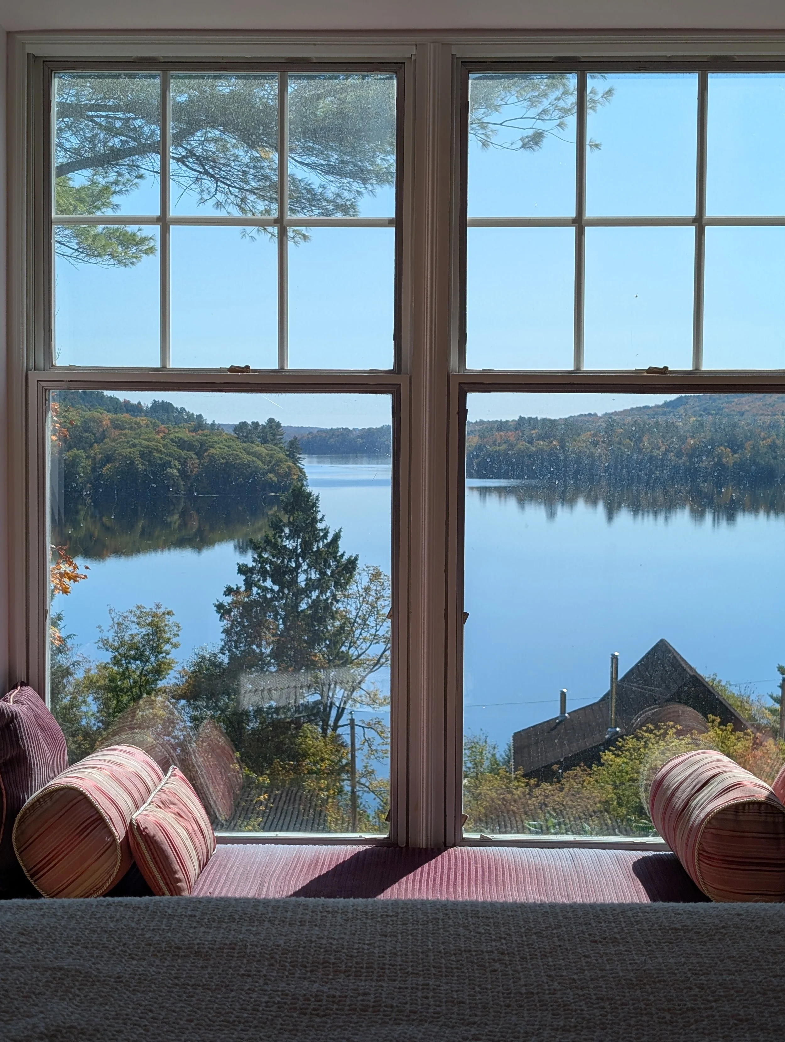 View through a window showing a peaceful lake surrounded by trees with fall foliage, from inside a room with a cushioned window seat and striped pillows.