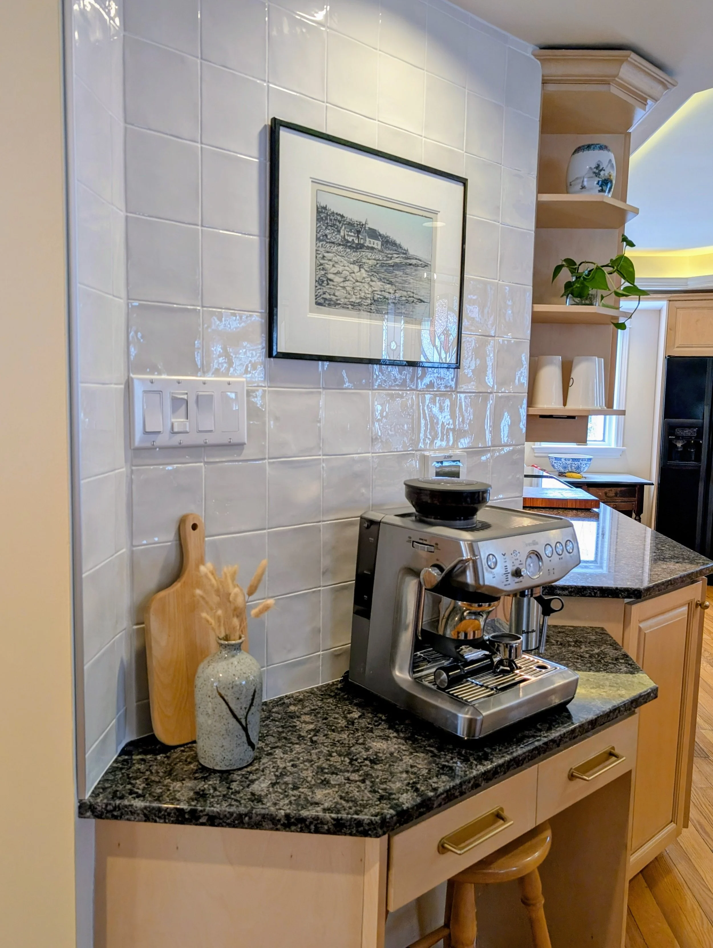 A kitchen countertop with a coffee machine, a cutting board, a vase with dried plants, a framed artwork of a landscape on white tiled wall, and open wooden shelves with decorative items and a potted plant.