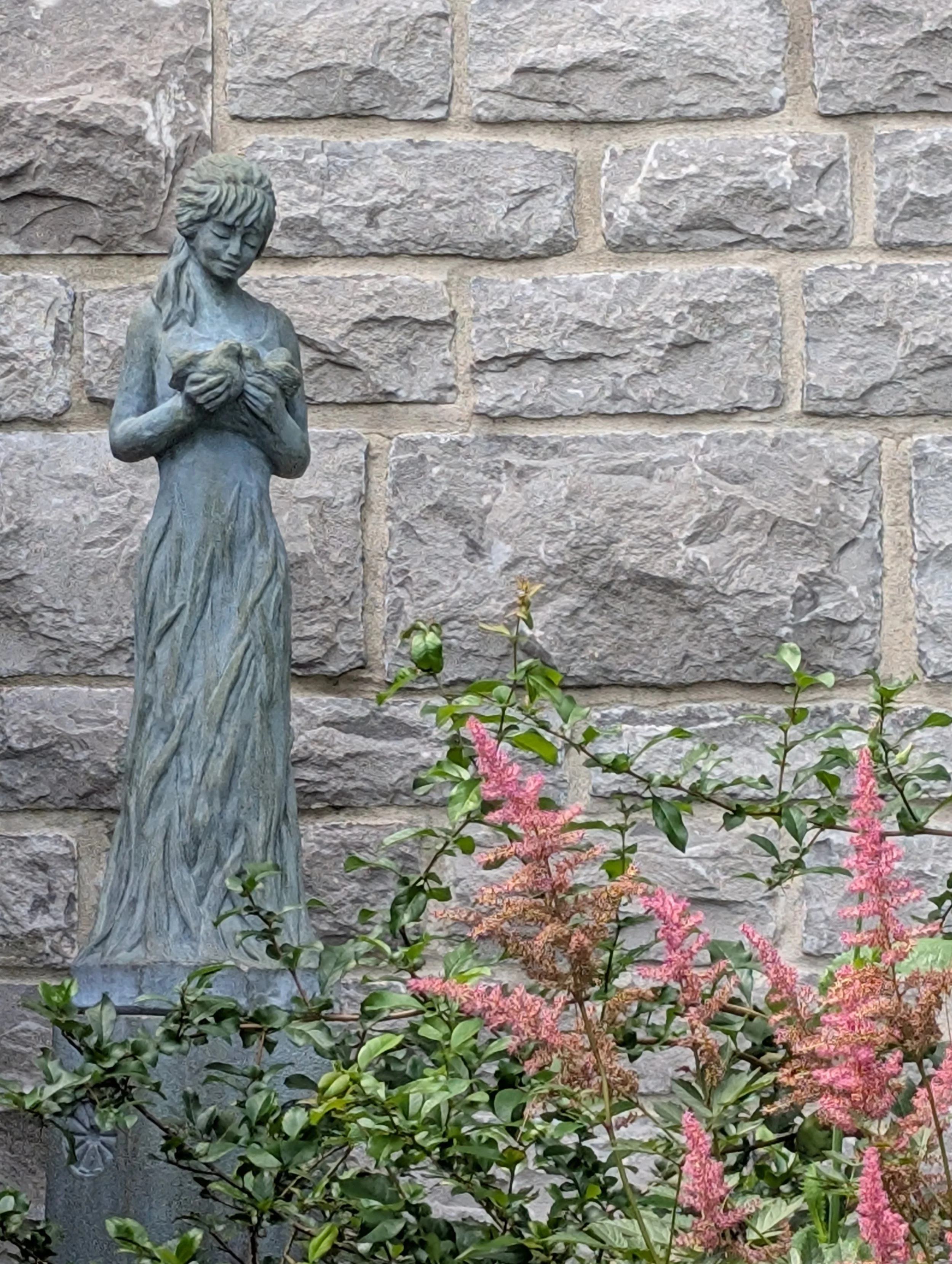 A statue of a woman holding a heart-shaped object, set against a stone wall with pink and green flowering plants in the foreground.
