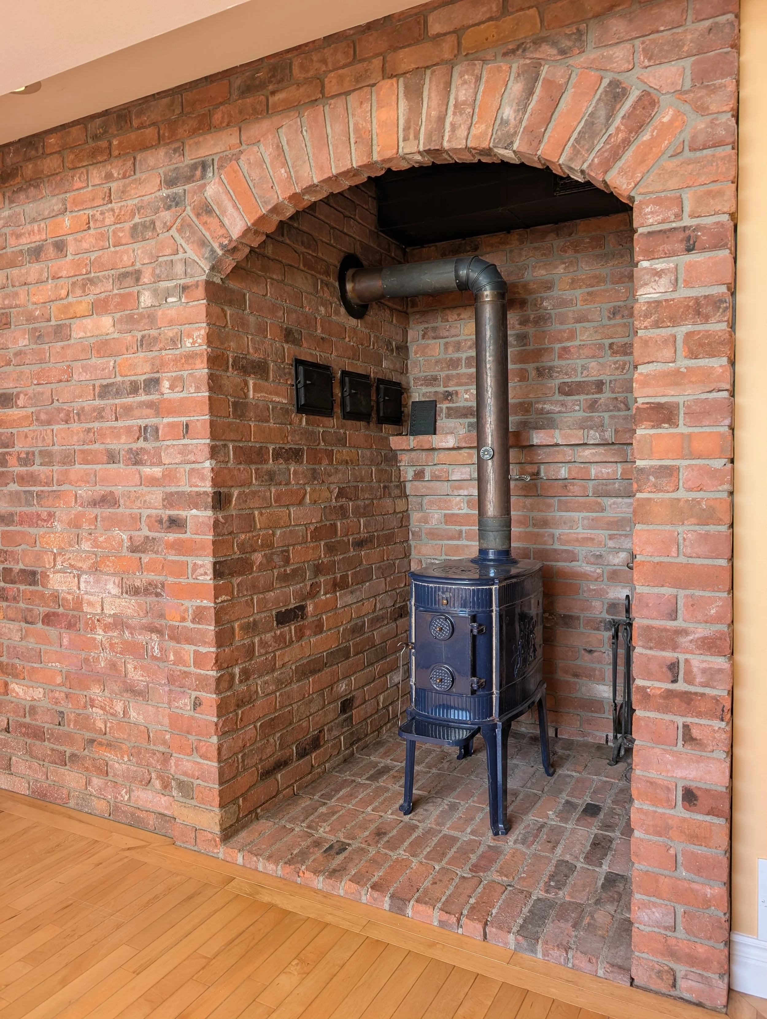 A vintage blue cast iron stove with a chimney pipe, set against a brick fireplace alcove with a raised brick hearth and three black metal vents on the wall beside it. The floor is light wood.