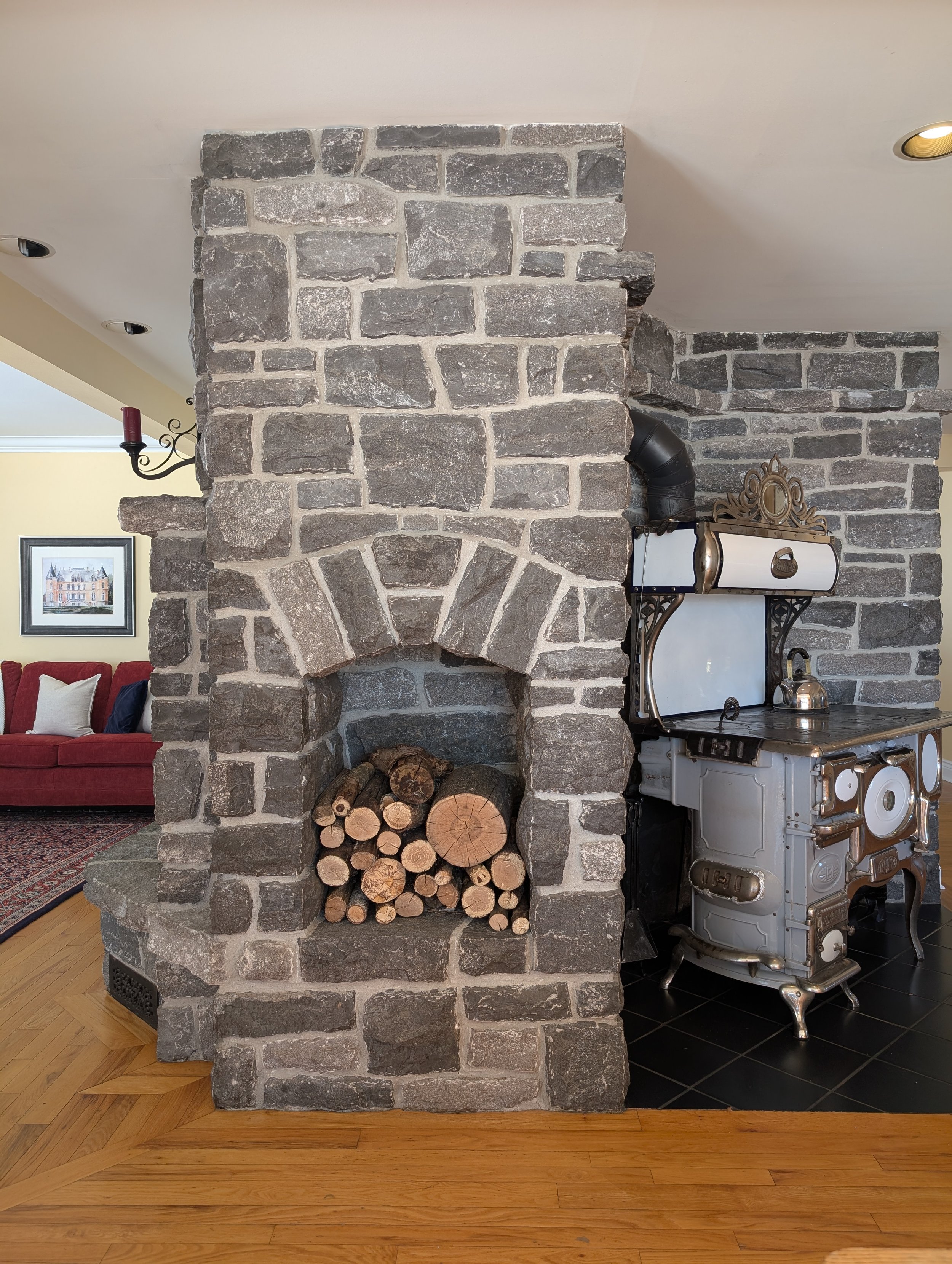 A room with a large stone fireplace filled with firewood, next to a vintage stove, and part of a living room with a red sofa and framed artwork on the wall.