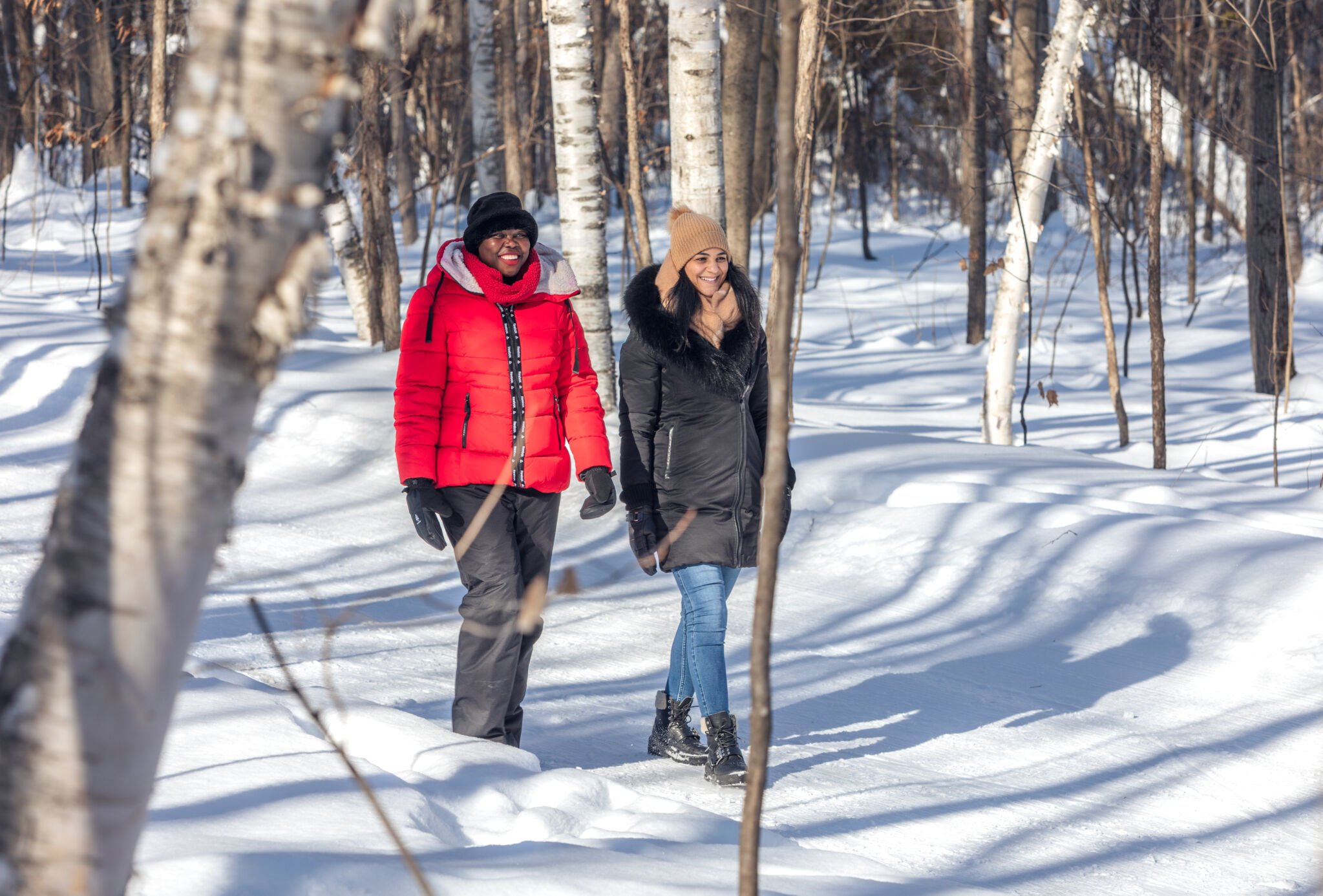 Two women walking through a snowy forest on a winter day, smiling and dressed in warm coats, hats, and gloves.